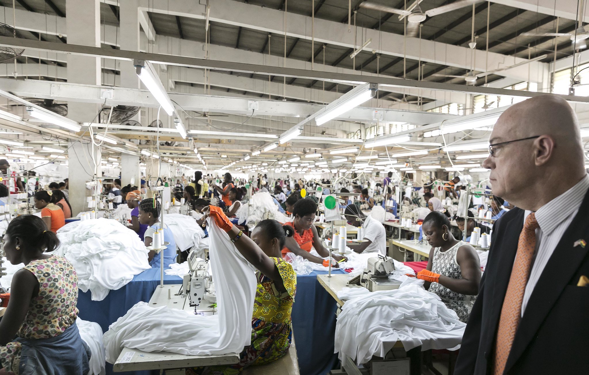 L'image montre une grande usine de couture animée. De nombreux ouvriers, principalement des femmes, sont occupés à coudre des vêtements, en particulier des chemises blanches. Ils travaillent en équipe autour de tables de couture, utilisant des machines à coudre. L'ambiance est dynamique, avec des mouvements rapides des mains et des tissus qui flottent. À droite, un homme en costume observe la scène, se tenant debout et faisant face au travail en cours. L'usine est bien éclairée, avec des lumières suspendues au plafond, et les murs sont de couleur claire, créant un espace de travail ouvert et organisé.