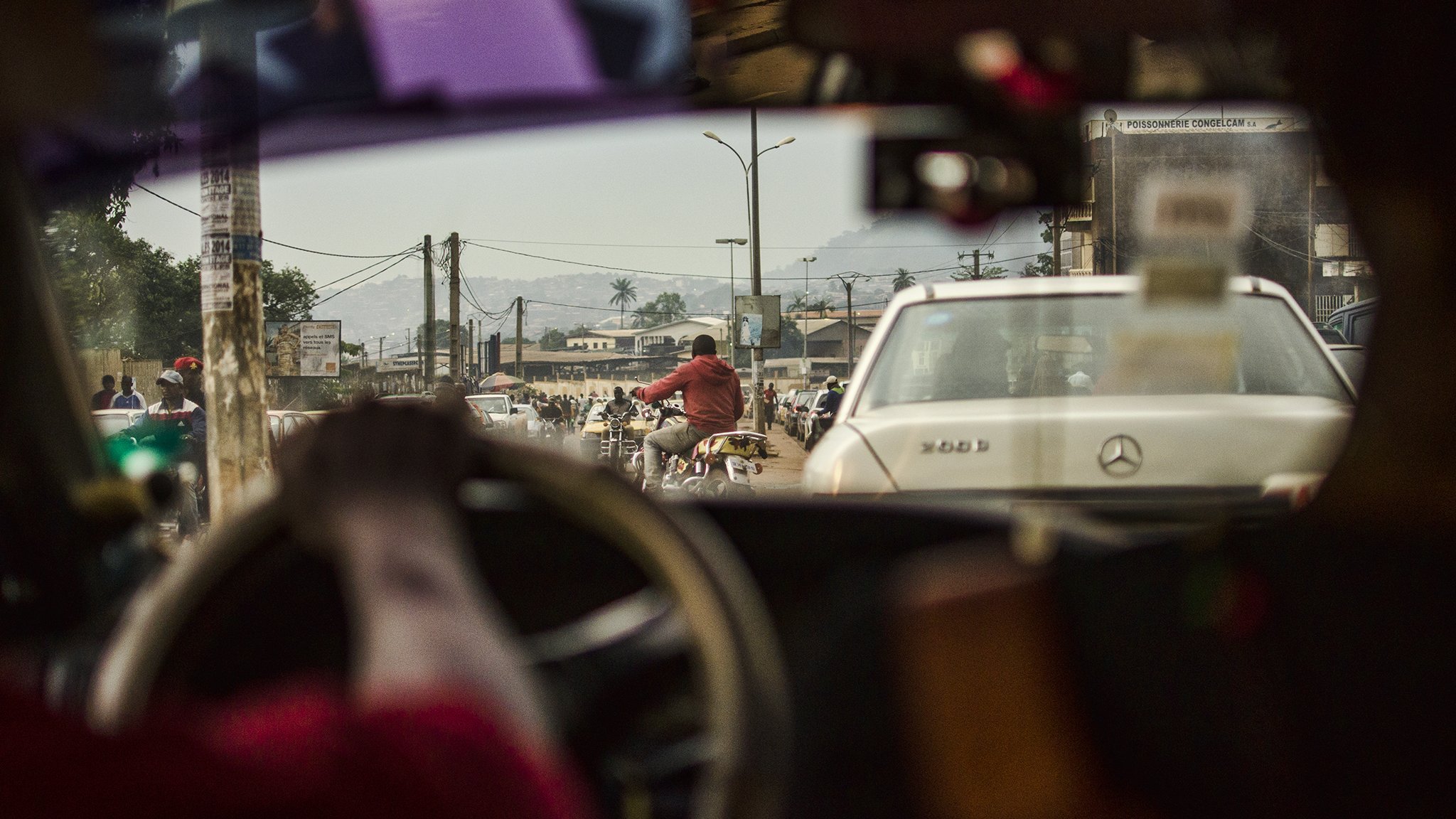 L'image présente une scène urbaine animée vue depuis l'intérieur d'un véhicule. On peut voir le côté d'un tableau de bord, avec une main tenant le volant. À travers le pare-brise, la circulation est dense, comprenant des voitures et des motos. On distingue une personne à vélo sur la route, et au fond, des bâtiments et des montagnes se dessinent à l'horizon. L'atmosphère semble vivante et mouvementée, caractéristique de la vie citadine. La lumière du jour éclaire la scène, révélant des détails sur les véhicules et les personnes qui circulent.