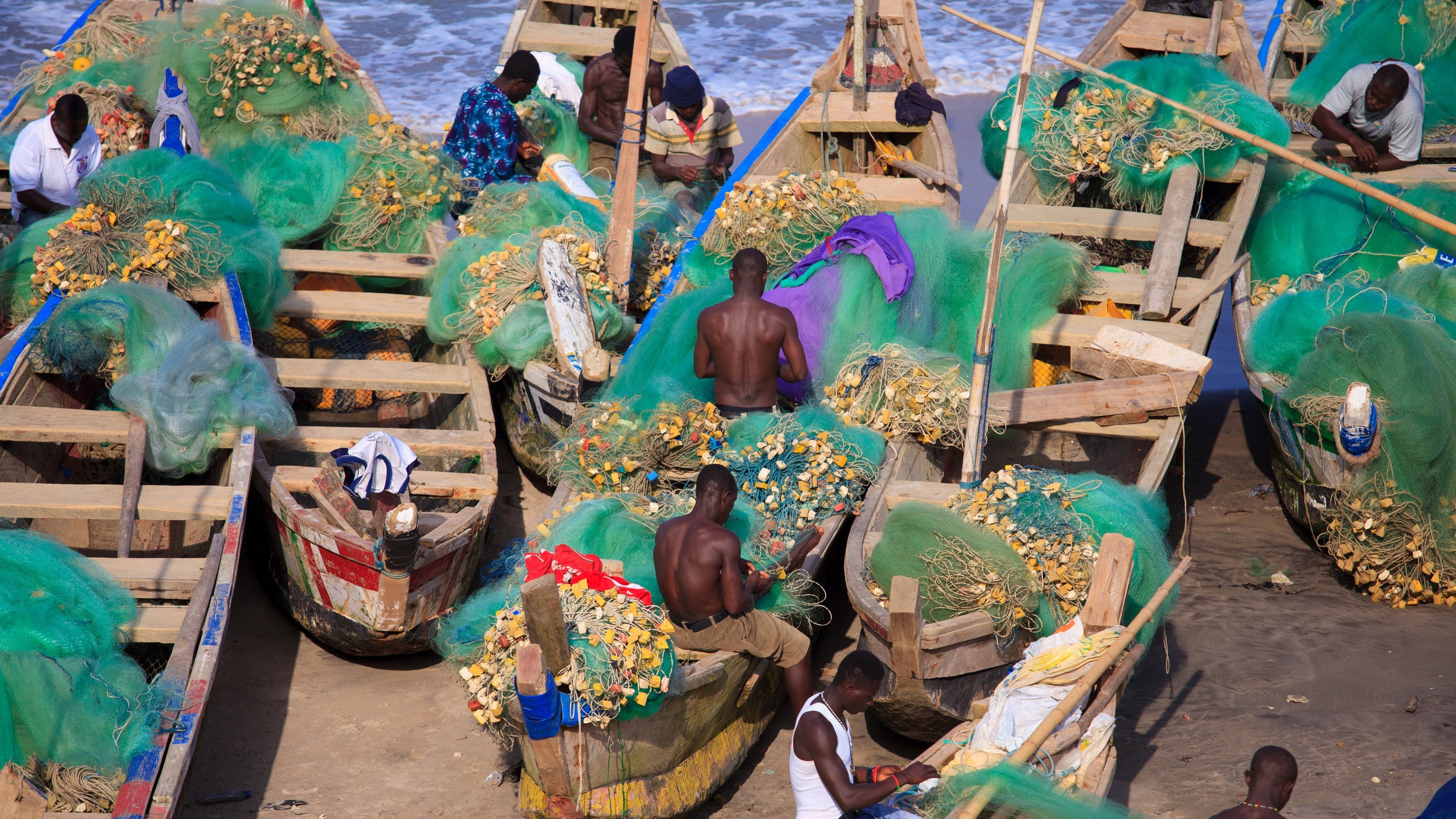 Cette image montre une scène vibrante en bord de mer, où plusieurs pêcheurs sont occupés à travailler sur des bateaux en bois. Les bateaux, peints de différentes couleurs, sont remplis de filets de pêche verts, ainsi que de coquillages et poisson. Les pêcheurs, principalement des hommes, sont torse nu et concentrés sur leurs tâches. L'arrière-plan révèle une mer calme avec des vagues douces, ajoutant une atmosphère de tranquillité à l'activité du port. L'air est imbibé de l'odeur de la mer et du bruit des vagues. Le soleil éclaire la scène, offrant une lumière chaude qui illumine le travail acharné des pêcheurs.