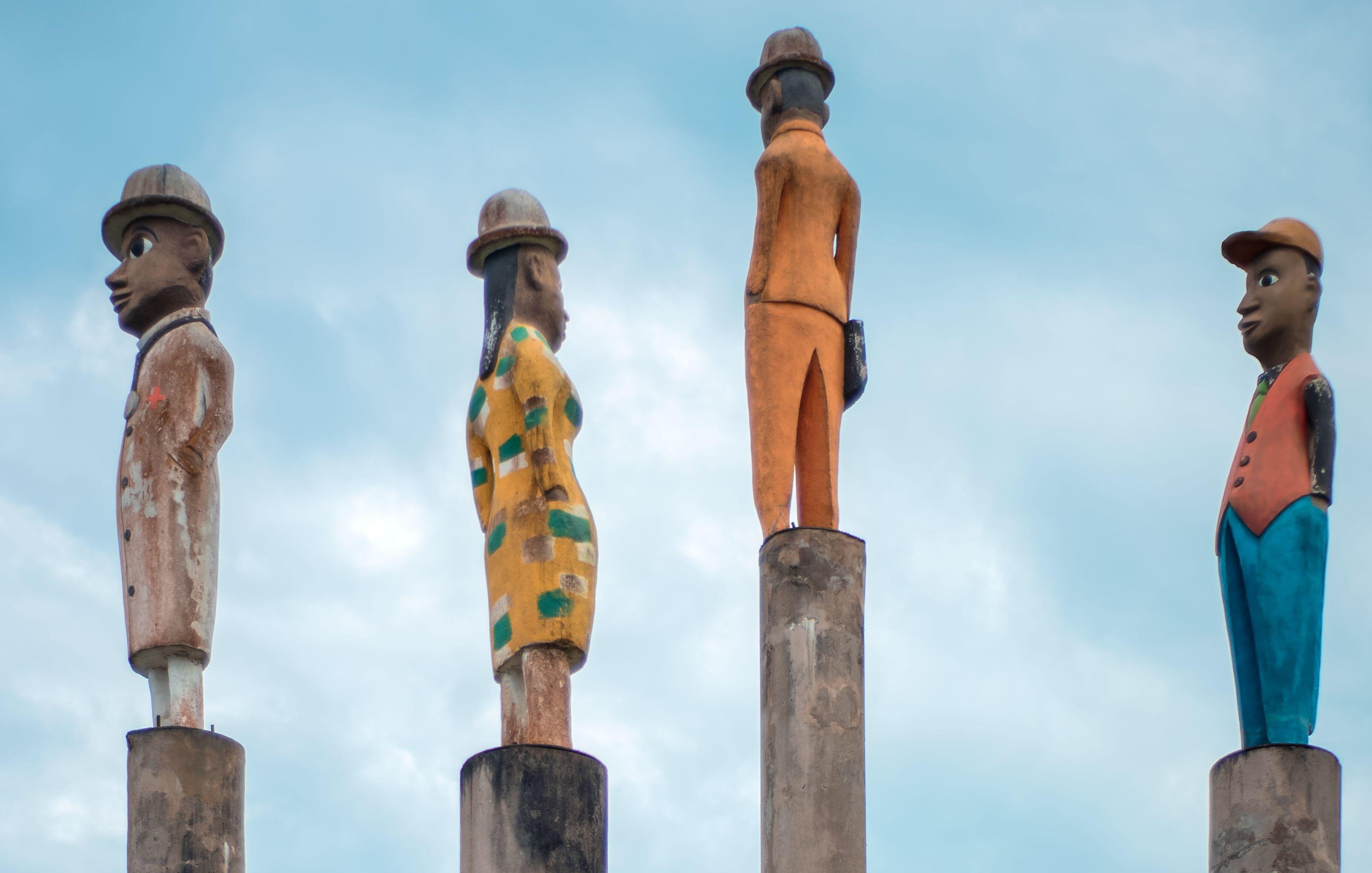 L'image présente quatre sculptures stylisées sur des colonnes. Chaque sculpture est peinte de manière différente, représentant des personnages humains : 1. La première sculpture à gauche est un homme avec un chapeau, vêtu d'un costume. Son expression est sérieuse. 2. La deuxième sculpture est une femme qui porte une robe avec un motif à rayures. Elle se tient droite, les bras le long du corps. 3. La troisième sculpture est un homme en orange, qui se tient également droit mais légèrement tourné sur le côté. 4. Enfin, la dernière sculpture est un homme en costume bleu, avec un chapeau, regardant vers l'horizon. L'arrière-plan est dominé par un ciel bleu clair, créant une ambiance paisible. Les sculptures semblent faire partie d'une installation artistique qui évoque la culture et l'identité. Les colonnes sur lesquelles elles reposent ajoutent une touche de hauteur et de grandeur à la scène.