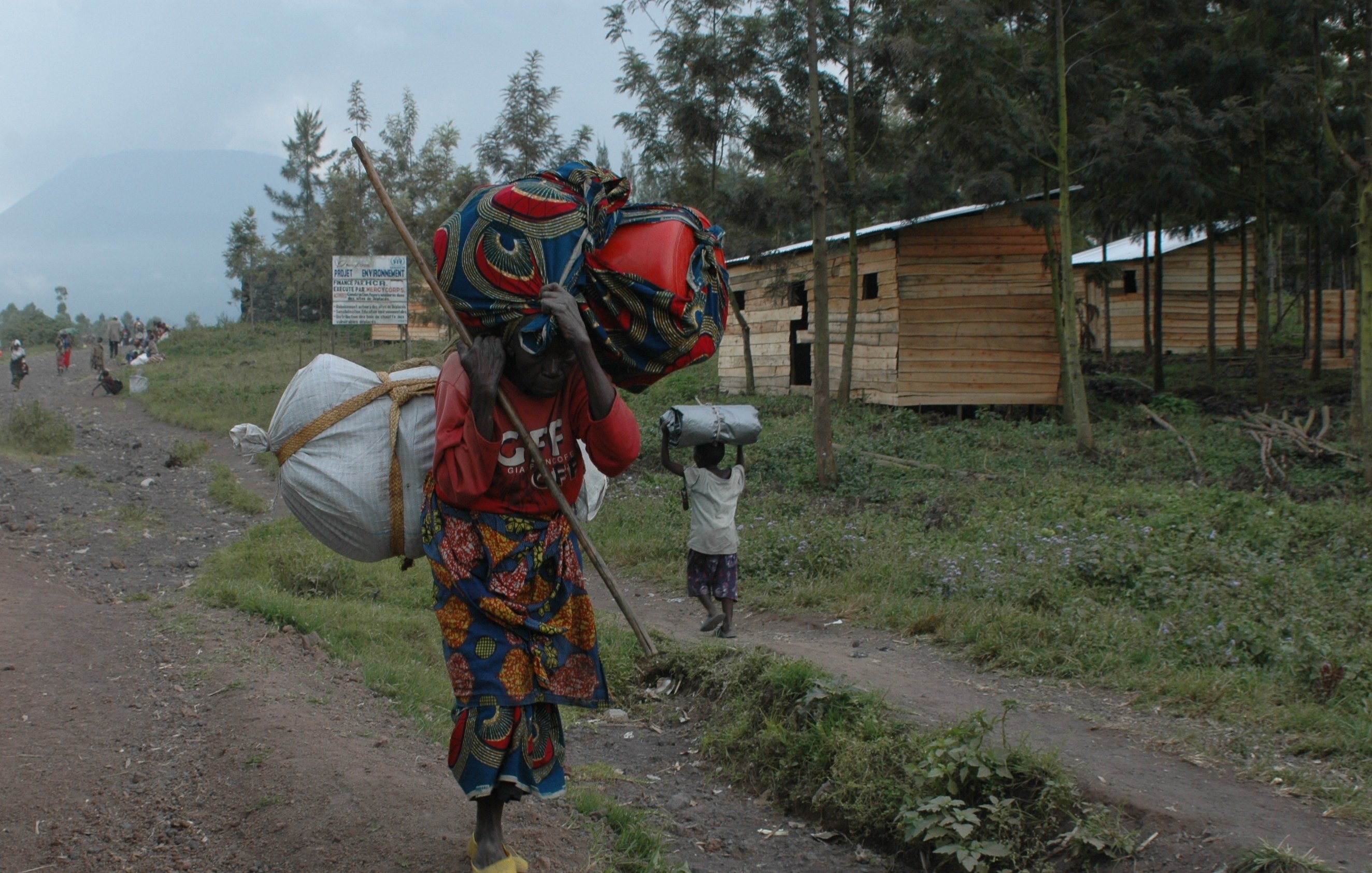 L'image montre un paysage rural d'une région montagneuse, probablement en Afrique. Sur la droite, vous voyez des maisons en bois, simples et fonctionnelles, avec un accent sur la nature environnante. À l'avant, une femme porte une charge lourde sur son dos, maintenue par un tissu coloré. Elle est vêtue d'une robe traditionnelle aux motifs vifs et colorés, qui contrastent avec le sol terreux et les feuillages verts. Dans le fond, une autre personne transporte également une charge, symbolisant la dureté du travail dans cette communauté. Les montagnes se dessinent en arrière-plan, offrant un sentiment d'immensité et de tranquillité, tandis que la scène évoque la résilience et le quotidien des habitants de cette région.
