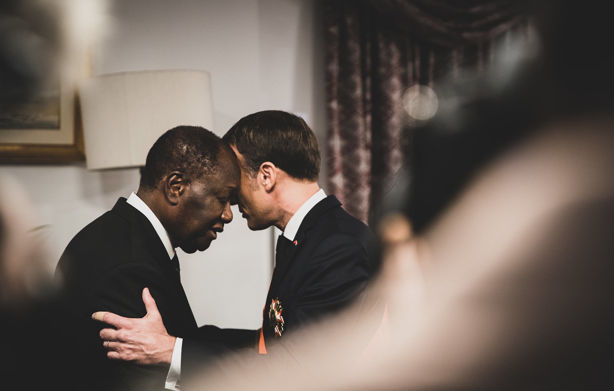 L'image montre deux hommes en train de se saluer de manière chaleureuse, leurs fronts se touchant, ce qui suggère une connexion profonde ou un moment d'émotion. L'un des hommes porte un costume sombre, tandis que l'autre semble également en habit formel. Le décor en arrière-plan, légèrement flou, indique un environnement intérieur, probablement un bureau ou une salle de conférences. La lumière est douce et crée une atmosphère respectueuse et intime. Les expressions des deux hommes semblent sérieuses et significatives, évoquant un échange important ou une reconnaissance mutuelle.