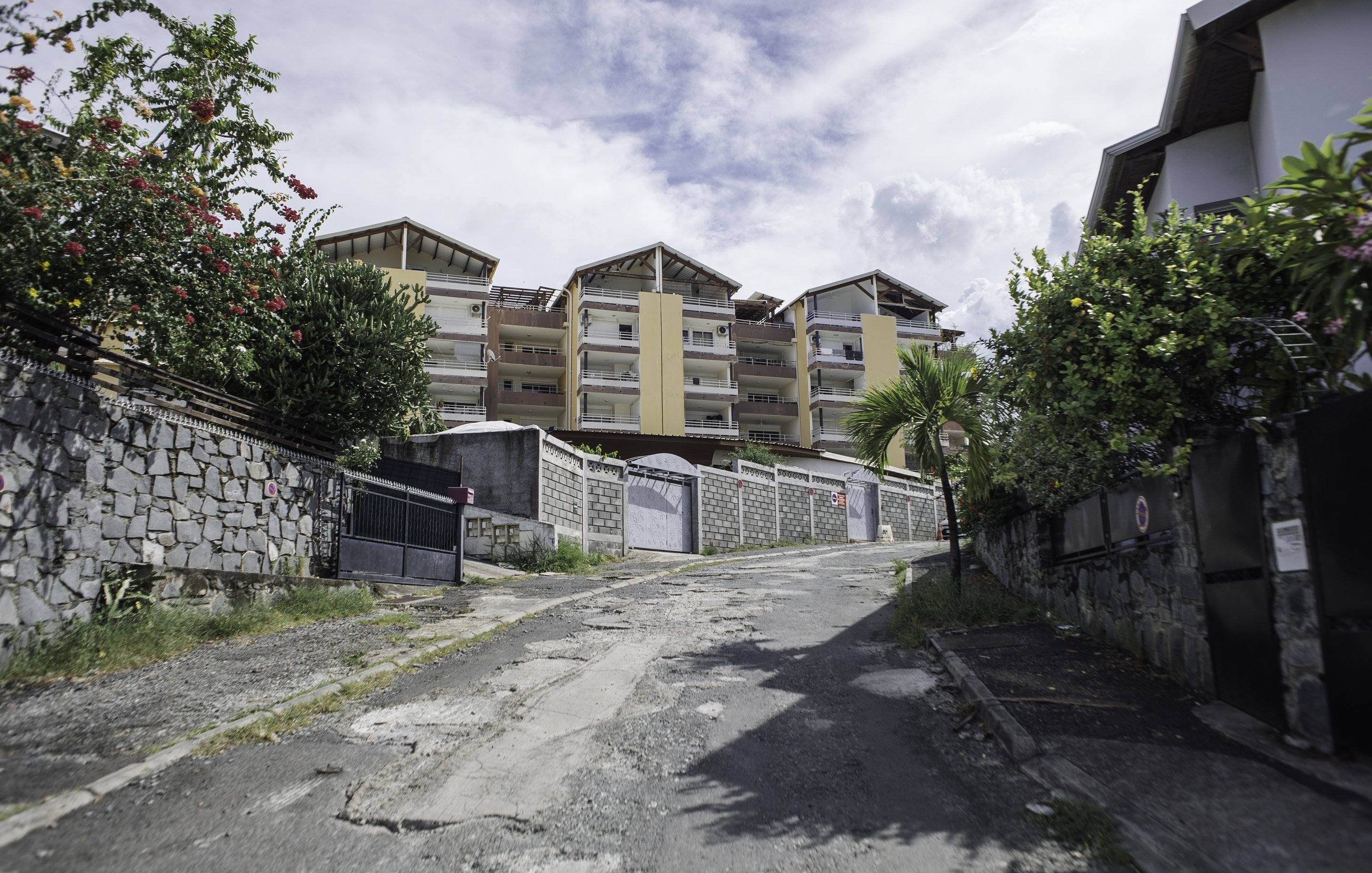 L'image montre une rue en pente, bordée de murs en pierre et de verdure. Sur le côté, on peut distinguer des fleurs colorées et des plantes tropicales. À l'arrière-plan, plusieurs bâtiments modernes avec des balcons se dressent, peints dans des teintes de jaune et de beige. Le ciel est nuageux, ce qui donne une ambiance légèrement dramatique à la scène. Le sol de la rue est inégal et montre des signes d'usure, avec des pavés qui apportent une texture variée au passage. L'atmosphère est calme, typique d'un quartier résidentiel.