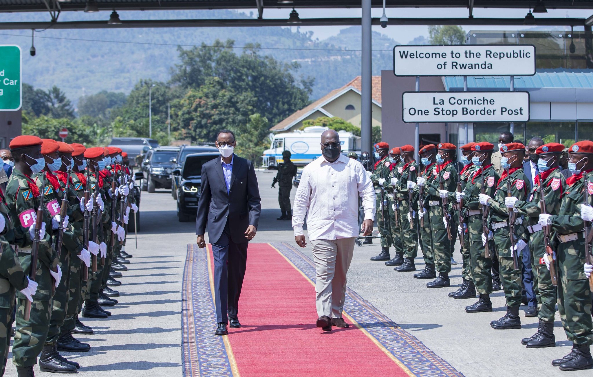 L'image montre un moment protocolaire à un poste frontalier. Deux hommes marchent sur un tapis rouge. À gauche, un homme en costume noir et masque, et à droite, un homme en chemise blanche. De chaque côté du tapis, des soldats en uniforme militaire attendent, formant une haie d'honneur. Les soldats portent des bérets rouges et des masques. En arrière-plan, on peut voir des panneaux indiquant la bienvenue au Rwanda et un poste frontière. L'ambiance est solennelle et cérémonielle.