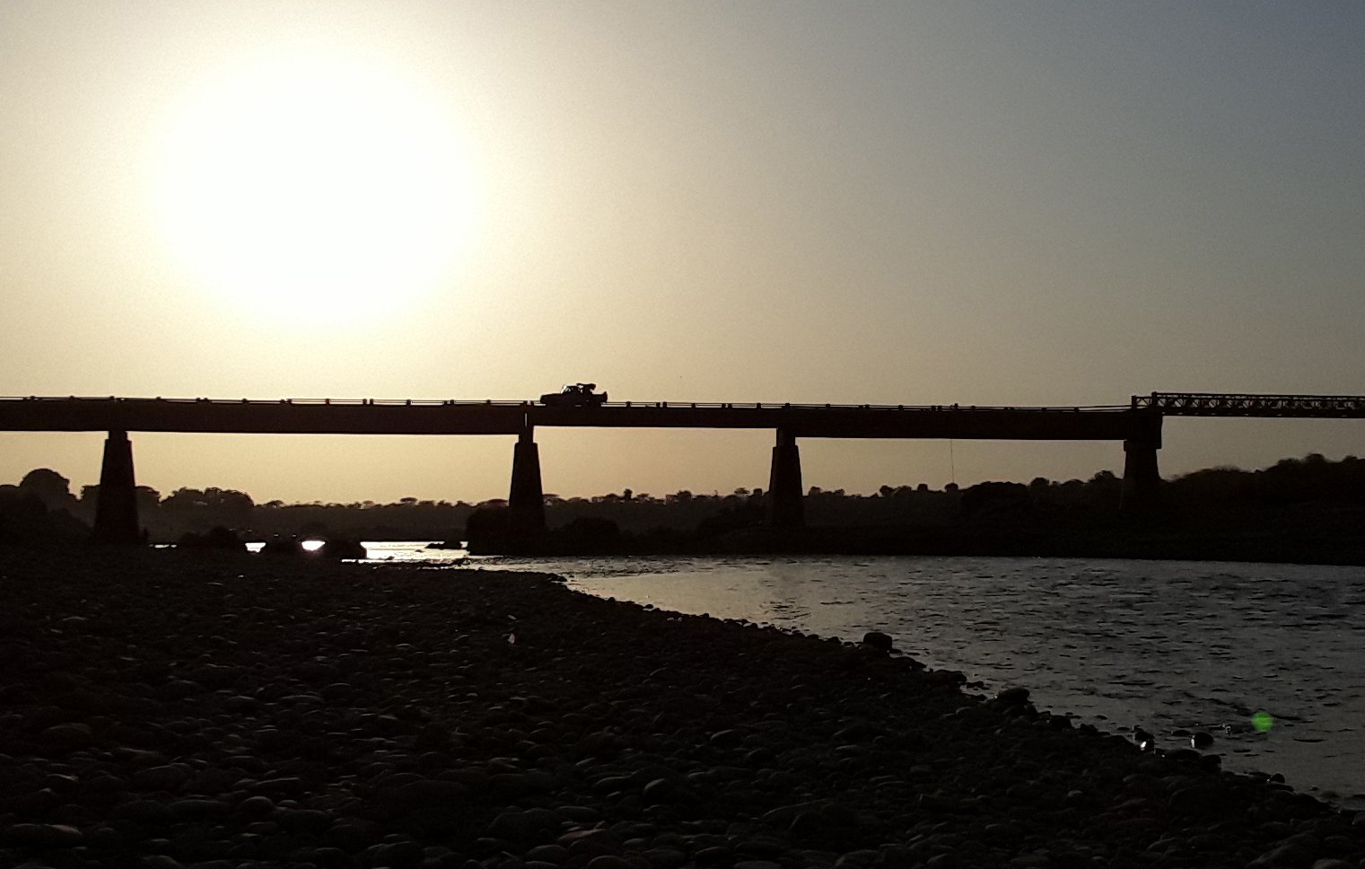 L'image montre un paysage côtier au crépuscule. À l'arrière-plan, on aperçoit un grand pont qui s'étend au-dessus d'une rivière. La lumière du soleil, basse à l'horizon, crée un contre-jour spectaculaire, rendant le pont et les éléments environnants en silhouette. Les contours de la structure sont sombres, se détachant des teintes dorées et oranges du ciel. À proximité de la rive, on peut imaginer des rochers ou des galets, ajoutant une texture au paysage. L'atmosphère est calme et paisible, évoquant un moment de tranquillité près de l'eau.