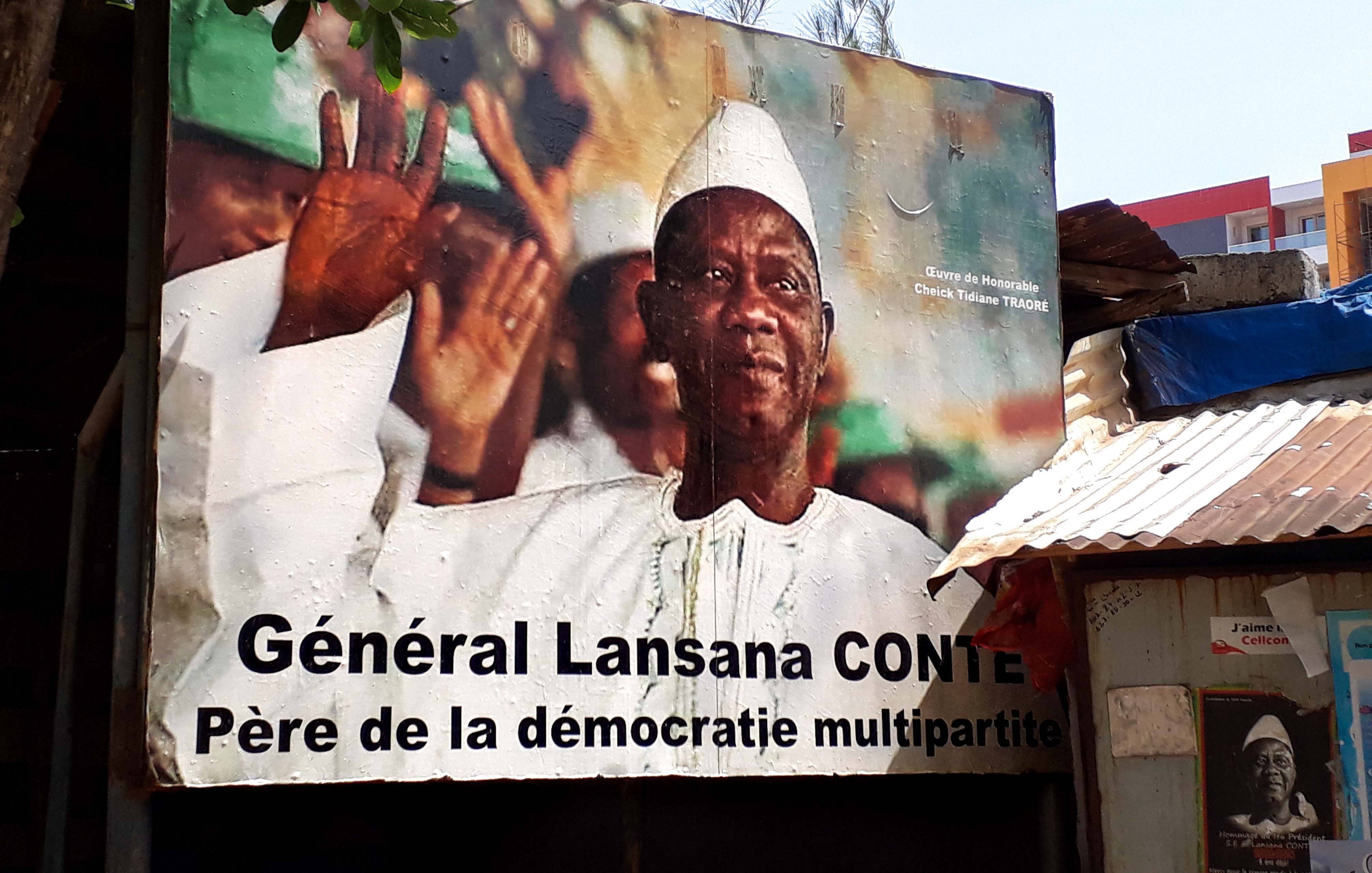 L'image représente une grande affiche murale d'un homme en train de saluer, avec un sourire bienveillant. Il porte une tenue traditionnelle, incluant un chapeau blanc et une chemise claire. En arrière-plan, on peut voir des couleurs vives qui semblent évoquer une atmosphère festive ou patriotique. Au bas de l'affiche, le texte indique « Général Lansana Conté, Père de la démocratie multipartite », soulignant ainsi son rôle important dans l'histoire politique. Les lignes et les formes des éléments de l'affiche créent un contraste qui attire le regard, suggérant un message de respect et d'honneur envers cette figure politique.