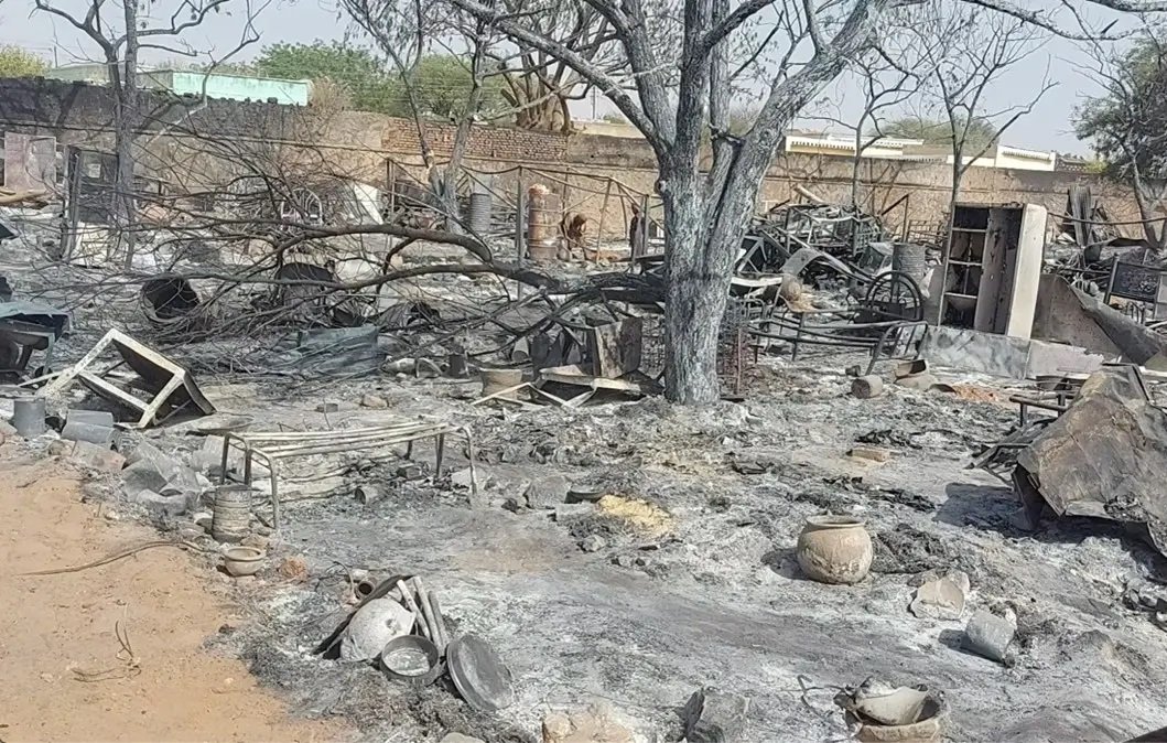 L'image montre un paysage de destruction et de désolation. Au premier plan, le sol est noirci par des cendres et des débris, témoignant d'un récent incendie. Des branches d'arbres calcinées s'élèvent vers le ciel, tandis que des objets métalliques, probablement des meubles ou des ustensiles, sont dispersés autour. On peut voir des pots brisés et des restes de ce qui semblait être une habitation ou un lieu de vie. L'atmosphère est sombre, reflétant la perte et la désolation causées par cet incident.