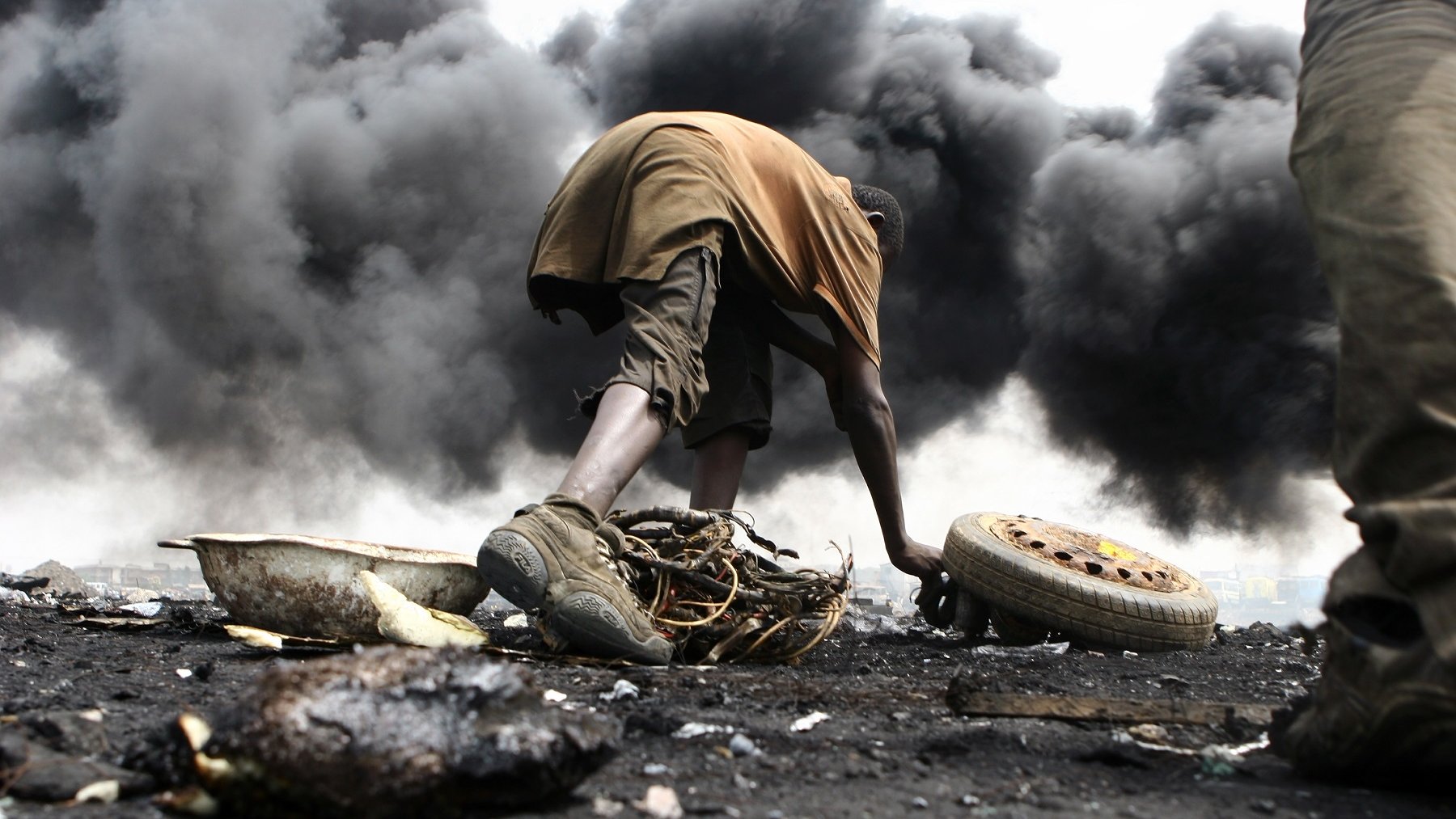 L'image montre un environnement très sombre et pollué, avec d'épaisses colonnes de fumée noire s'élevant dans l'air. Au premier plan, un jeune homme est accroupi, en train de manipuler des débris. Il porte des vêtements simples, à moitié sales, et semble concentré sur ce qu'il fait. Son environnement est chaotique, avec des morceaux de métal, de la ferraille et des pneus éparpillés autour de lui. La scène dégage une atmosphère de difficulté et de lutte, illustrant les conditions de vie dans cet endroit touché par la pollution et la dégradation environnementale.