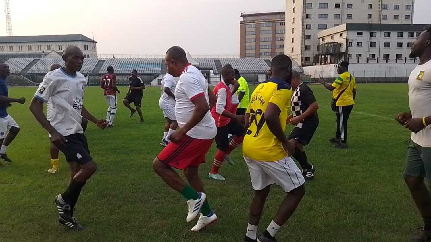L'image représente un groupe de personnes jouant au football sur un terrain en herbe. Les joueurs portent des maillots de différentes couleurs, principalement blancs, rouges et jaunes. On peut voir des hommes en train de s'échauffer ou de danser, avec des mouvements dynamiques et joyeux. En arrière-plan, on aperçoit des gradins remplis, et des bâtiments se dressent autour du terrain. L'ambiance semble conviviale et énergique, avec un sentiment de camaraderie parmi les joueurs.