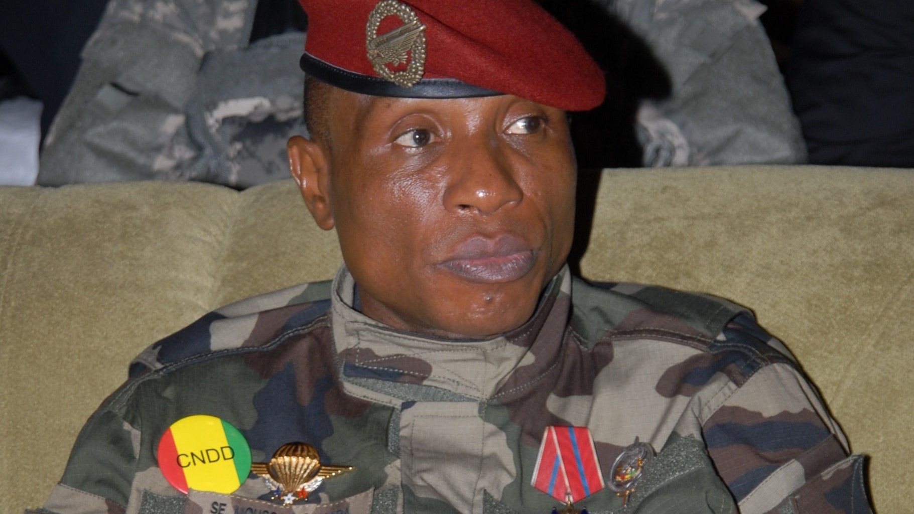 L'image montre un militaire assis, vêtu d'un uniforme camouflage. Il porte une casquette rouge ornée d'un insigne. Sur sa poitrine, il y a plusieurs décorations et médailles visibles. Les couleurs de l'uniforme et des insignes, notamment le jaune et le rouge, évoquent des éléments de son pays. Son expression est sérieuse, et il semble attentif, probablement à une discussion ou à un événement. L'arrière-plan est flou, suggérant une atmosphère formelle ou solennelle.