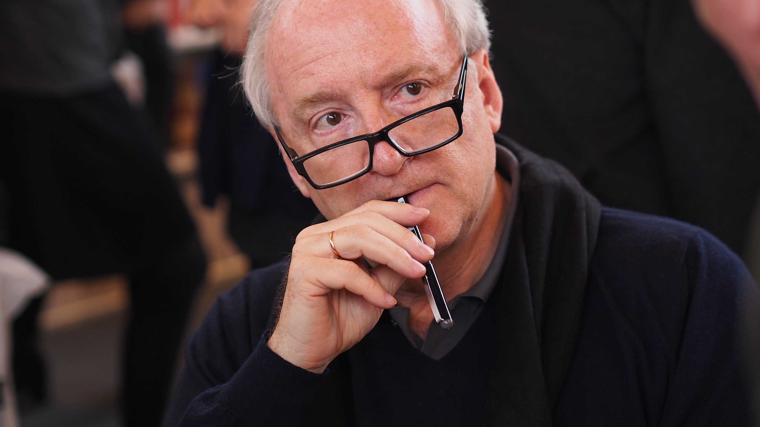 L'image montre un homme assis, vêtu d'un pull sombre, avec des lunettes sur le nez. Il porte une expression pensive, tenant un stylo près de sa bouche, comme s'il était en train de réfléchir ou de considérer quelque chose de manière approfondie. En arrière-plan, on distingue d'autres personnes floues, suggérant une ambiance de discussion ou d'échange, typique d'un événement de rencontre ou de signature de livres. La lumière est douce, créant une atmosphère chaleureuse.