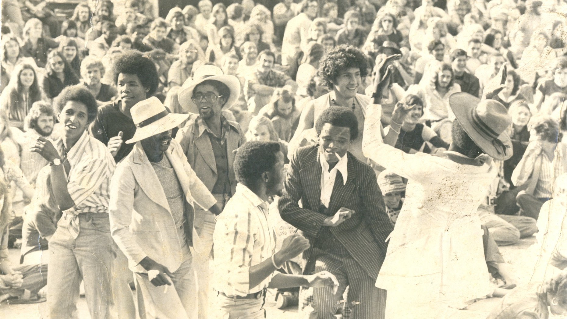 L'image représente une scène vibrante de fête en plein air, probablement lors d'un concert ou d'un festival. Au premier plan, un groupe de personnes danse joyeusement, exprimant de l'énergie et de la convivialité. Les danseurs, de différentes origines, portent des vêtements variés, allant des costumes élégants à des tenues plus décontractées. En arrière-plan, une foule dense assiste à la scène, créant une ambiance de camaraderie. Les visages sont souriants, captivés par la musique et l'atmosphère festive. Le tout suggère une célébration de la vie et de la culture, où la danse et la communion entre les gens sont à l'honneur. L'éclairage semble naturel, évoquant une belle journée ensoleillée.