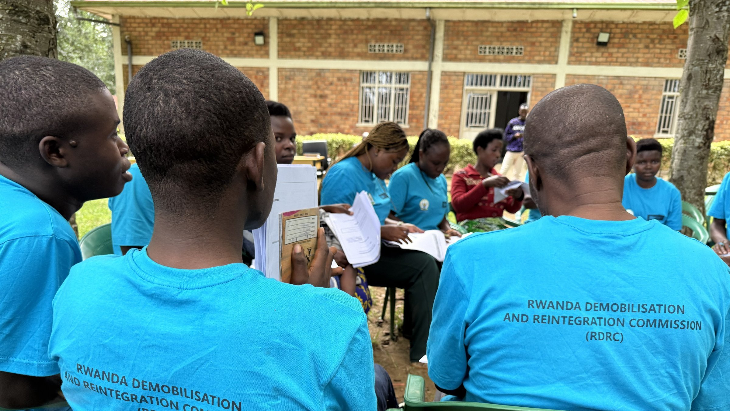 L'image montre un groupe de personnes assises à l'extérieur. Elles portent toutes des t-shirts bleu clair avec le texte "RWANDA DEMOBILISATION AND REINTEGRATION COMMISSION (RDRC)" imprimé au dos. On peut apercevoir des chaises en plastique vertes sur lesquelles elles sont assises. À l'arrière-plan, plusieurs personnes sont en train de discuter, et un bâtiment en briques apparaît légèrement flou derrière eux. L'ambiance semble conviviale et engagée, avec des participants qui semblent suivre une activité ou une formation en lien avec leur programme.