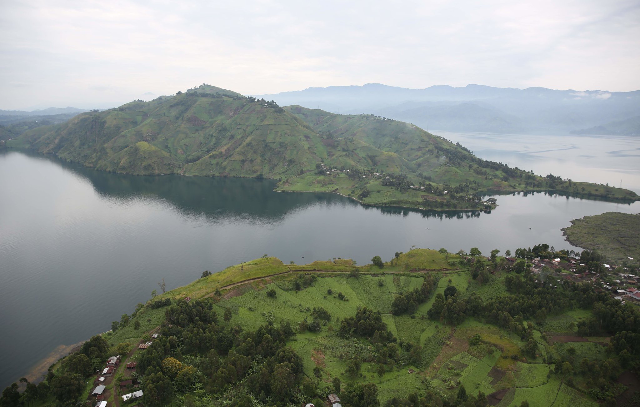 Imaginez un vaste paysage naturel serein, où les collines verdoyantes et les rives d'un lac s'étendent à perte de vue. Au premier plan, des champs verdoyants semblent onduler sur le sol, parsemés de petites habitations rustiques. En arrière-plan, une immense colline se dresse majestueusement, recouverte de nuances de vert qui varient avec la lumière. Le lac, calme et réfléctif, capture le ciel gris clair au-dessus. À l'horizon, d'autres collines se dessinent, créant une impression de profondeur et de tranquillité dans ce cadre apaisant. L'ensemble dégage une atmosphère de paix et de beauté naturelle, accentuée par le murmure léger de l'eau.