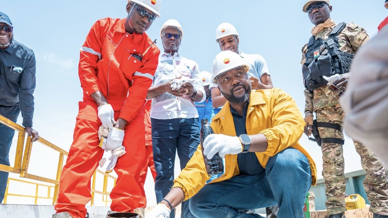 Dans cette image, on voit des hommes en tenue de travail sur une plateforme en extérieur. Deux d'entre eux portent des casques de sécurité blancs et des vêtements de protection, l'un en orange et l'autre en jaune. Le premier, en orange, se penche pour examiner un récipient, tandis que le second, en jaune, est à genoux et tient un échantillon de liquide dans une bouteille. En arrière-plan, d'autres personnes regardent, vêtues de manière décontractée. Le ciel est dégagé, suggérant une journée ensoleillée. L'environnement semble être lié à un travail industriel ou d'ingénierie, avec des installations visibles autour de eux.