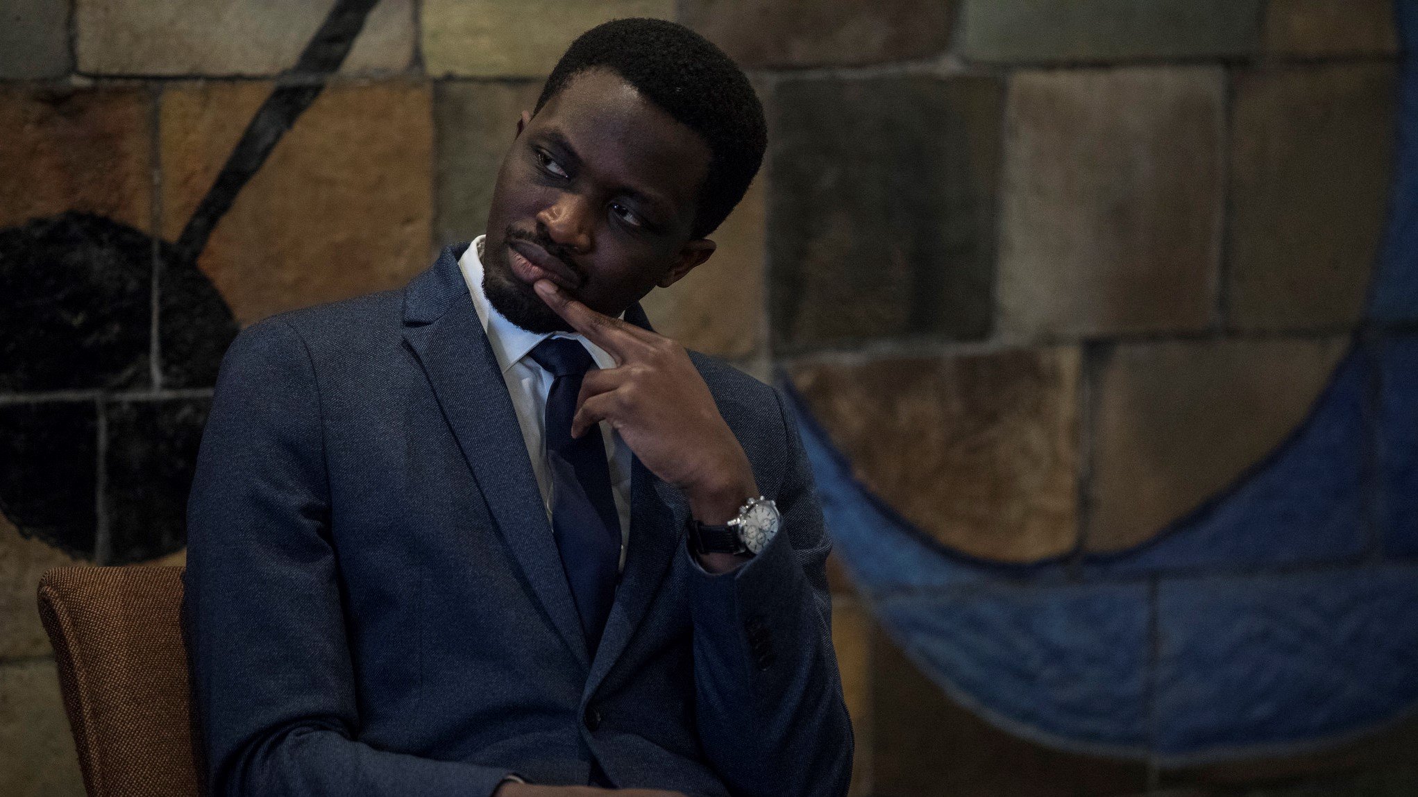 L'image montre un homme assis dans un environnement intérieur. Il porte un costume sombre avec une chemise claire et une cravate, ce qui dénote un certain formalisme. Son expression est pensive, avec un doigt sur le menton, suggérant qu'il réfléchit à quelque chose de sérieux. En arrière-plan, on aperçoit un mur aux motifs colorés, avec des formes géométriques qui ajoutent une touche artistique à la scène. L'ambiance est calme et réfléchie.