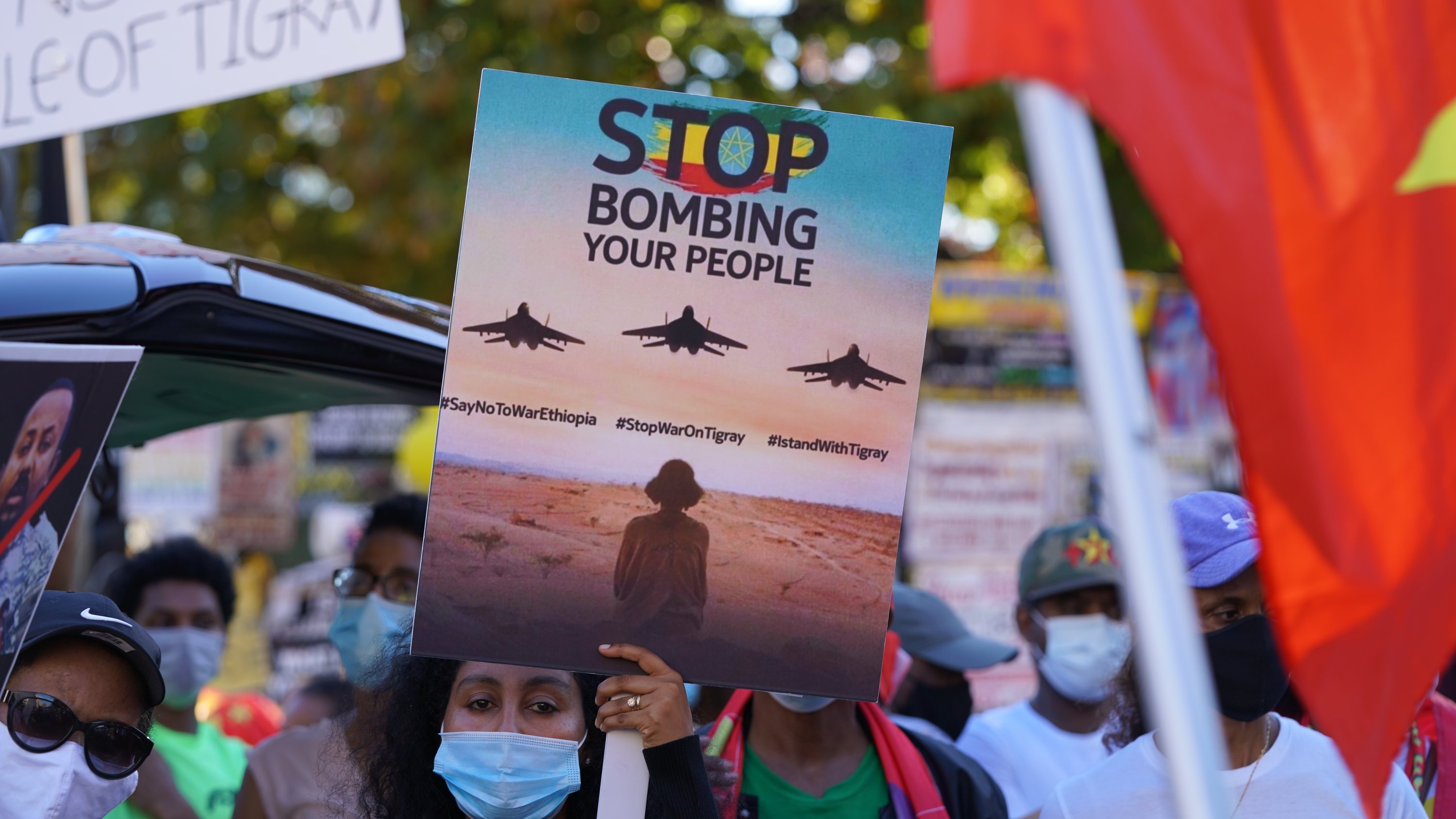 L'image montre une scène de manifestation. Au premier plan, une personne tient une grande pancarte qui affiche le message "STOP BOMBING YOUR PEOPLE" avec des images d'avions de chasse en vol. La personne est entourée d'autres manifestants portant des masques faciaux, certains brandissant d'autres pancartes. En arrière-plan, on peut voir des drapeaux et des affiches qui expriment des messages de protestation. L'atmosphère est chargée d'émotion, soulignant un appel urgent à l'action.