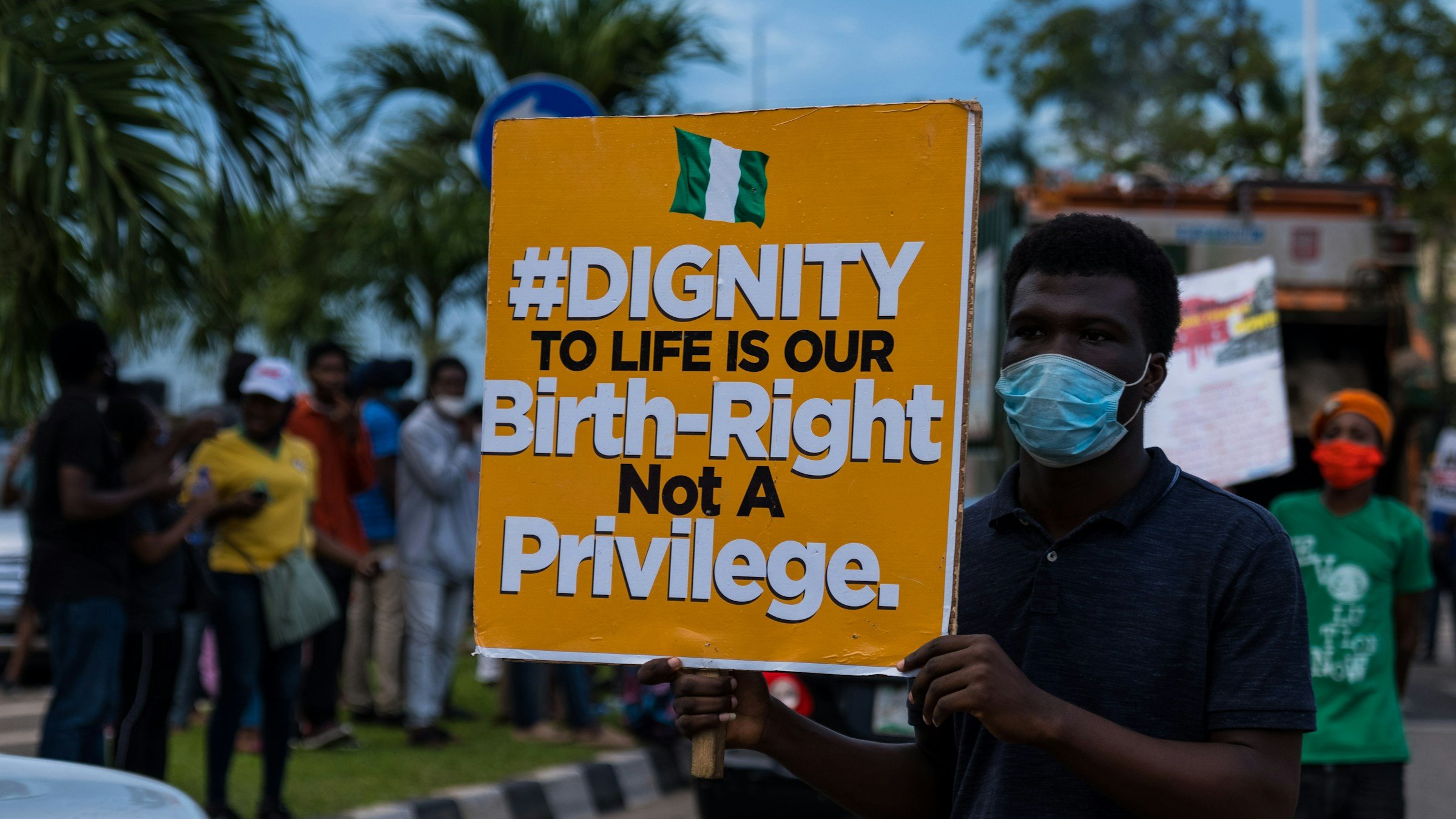Sur l'image, une manifestation a lieu dans la rue. Un homme se tient au premier plan, portant un masque. Il tient une grande pancarte colorée qui affiche le message suivant : "#DIGNITY TO LIFE IS OUR Birth-Right Not A Privilege" en lettres grandes et noires sur un fond orange. En arrière-plan, on aperçoit un groupe de personnes, certaines portant également des masques, rassemblées autour de lui. L'environnement est urbain, avec des arbres et des véhicules visibles. L'atmosphère est engagée, témoignant d'une lutte pour les droits et la dignité humaine.