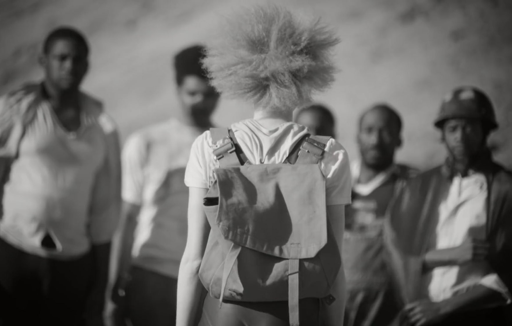 L'image présente un groupe de personnes, avec au premier plan une personne aux cheveux crépus et blonds, vue de dos. Elle porte un sac à dos. L'arrière-plan est flou, mais on devine des montagnes ou des collines. Les autres individus, qui se tiennent plus loin, ont des expressions faciales sérieuses et semblent réunis dans une conversation ou une interaction. L'atmosphère de l'image est en noir et blanc, ce qui accentue les contrastes entre les différentes textures et les attitudes des personnages.