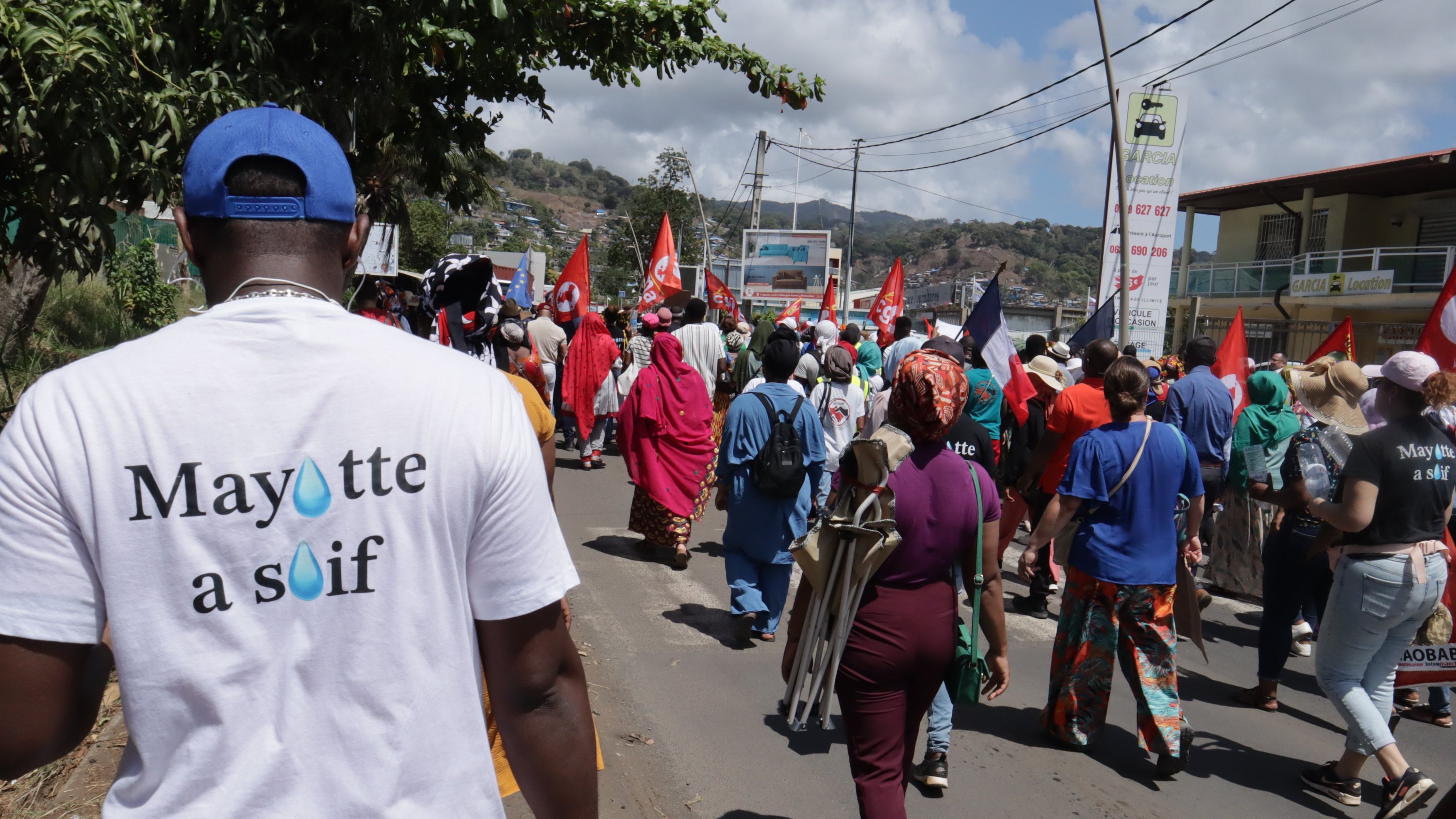 La scène dépeint un rassemblement animé dans une rue, où un groupe de manifestants avance. Au premier plan, on aperçoit une personne portant un t-shirt blanc avec l'inscription "Mayotte à souff" sur le dos. Les manifestants, vêtus de couleurs variées, portent des drapeaux rouges et d'autres pancartes, créant une atmosphère de solidarité et de revendication. À l'arrière-plan, on voit des bâtiments et des montagnes, sous un ciel partiellement nuageux. Les sons de la foule, les discussions et les chants de protestation résonnent, ajoutant à l'énergie de l'événement.