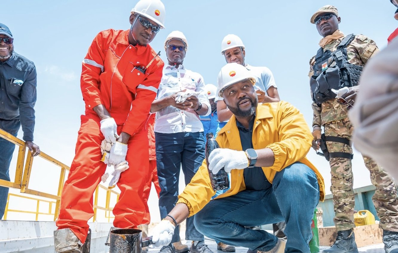 Dans cette image, on voit des hommes en tenue de travail sur une plateforme en extérieur. Deux d'entre eux portent des casques de sécurité blancs et des vêtements de protection, l'un en orange et l'autre en jaune. Le premier, en orange, se penche pour examiner un récipient, tandis que le second, en jaune, est à genoux et tient un échantillon de liquide dans une bouteille. En arrière-plan, d'autres personnes regardent, vêtues de manière décontractée. Le ciel est dégagé, suggérant une journée ensoleillée. L'environnement semble être lié à un travail industriel ou d'ingénierie, avec des installations visibles autour de eux.