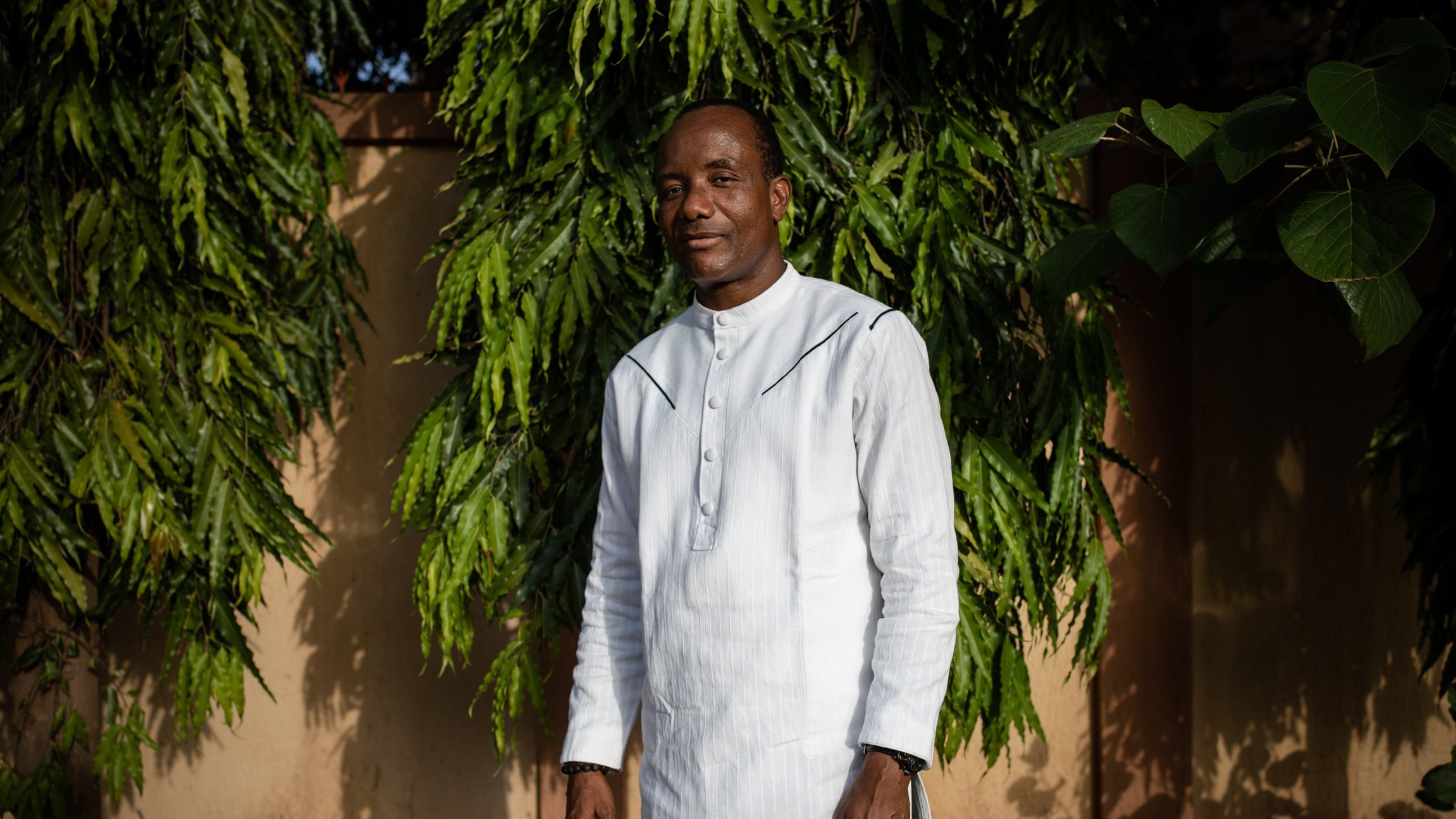 L'image montre un homme debout devant un arrière-plan de feuillage vert dense. Il porte une tunique blanche à col montant et un pantalon sombre. Son sourire dégage une attitude amicale et confiante. Les arbres autour de lui ajoutent une ambiance naturelle, avec des branches feuillues qui encadrent sa silhouette. Le fond est constitué de murs dans des tons nude, apportant une touche chaleureuse à la scène. La lumière naturelle semble douce, créant une atmosphère sereine.