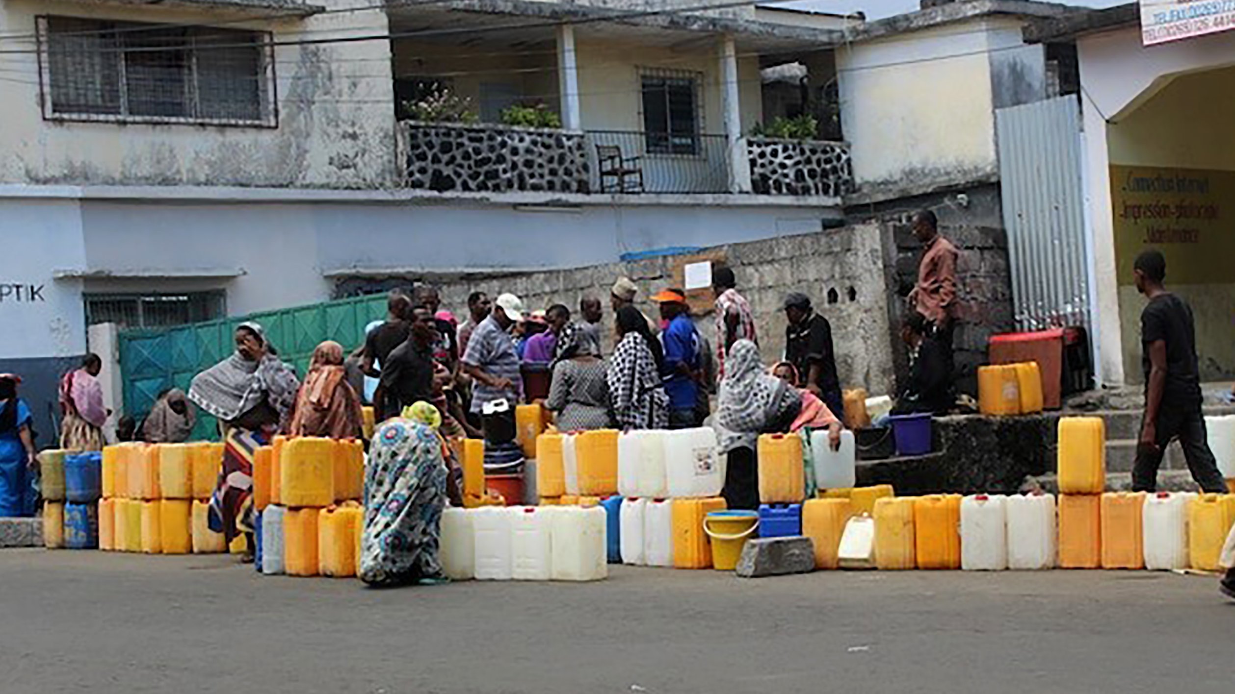 L'image montre une scène animée dans une rue. Des groupes de personnes se rassemblent autour de plusieurs conteneurs en plastique de différentes tailles, souvent de couleur orange ou jaune. Ces conteneurs sont alignés sur le sol. Les personnes, principalement habillées de vêtements traditionnels, semblent attendre leur tour pour remplir les récipients d'eau. En arrière-plan, on distingue des bâtiments aux façades simples, certains avec des balcons. L'ambiance est celle d'une activité quotidienne, soulignant l'importance de l'eau dans la vie des gens.