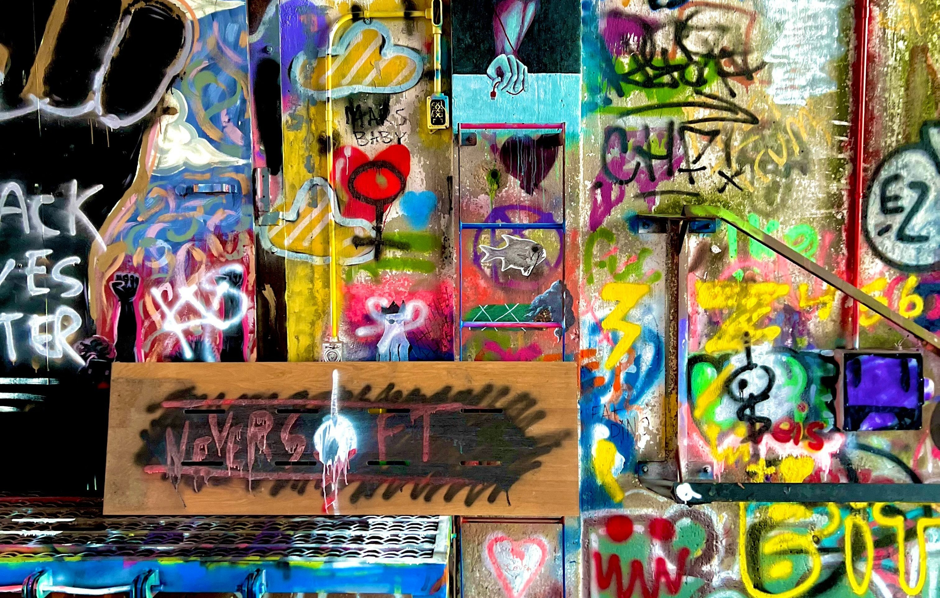 Cette image montre un mur vibrant de graffiti et d'art urbain. Les couleurs éclatantes, comme le jaune, le rouge, le bleu et le vert, créent un tableau chaotique et dynamique. On peut y voir des symboles et des mots, notamment un poing levé, représentant la résistance, ainsi que des cœurs et des dessins abstraits. Une planche en bois au centre affiche des mots en noir, évoquant des thèmes de persévérance. L'ensemble dégage une atmosphère de créativité libre et d'expression personnelle, transformant un espace ordinaire en une œuvre d'art collective.