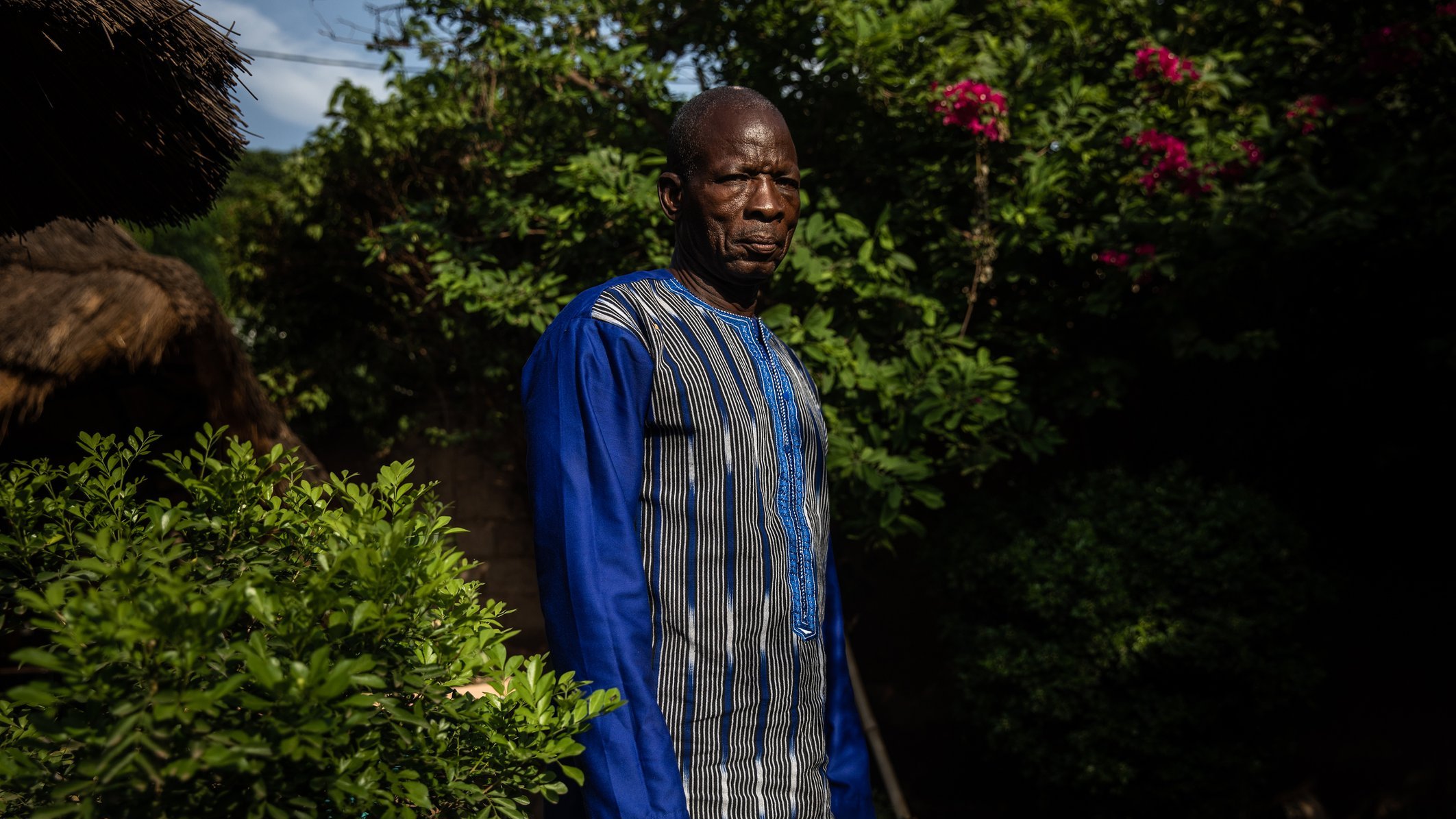 L'image montre un homme debout dans un environnement verdoyant. Il porte une tunique à manches longues, qui est ornée de lignes bleues et de motifs subtils. Son expression est sérieuse et son regard est dirigé vers l'objectif. Autour de lui, on peut apercevoir des plantes luxuriantes, avec des feuilles brillantes et quelques fleurs colorées en arrière-plan. La lumière du soleil filtre à travers les branches, créant une atmosphère tranquille.