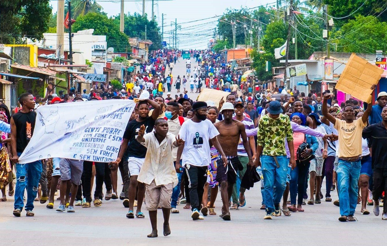 L'image montre une foule nombreuse marchant dans une rue animée. Les gens sont regroupés, certains tiennent des banderoles ou des pancartes. L'ambiance semble dynamique, avec beaucoup de mouvements et des échanges d'expressions entre les participants. On peut également ressentir une diversité dans les vêtements, allant des tenues décontractées aux vêtements plus colorés. En arrière-plan, des boutiques et des bâtiments bordent la rue, tandis que des fils électriques sont suspendus au-dessus. Les voix et les chants des manifestants semblent animer l'espace, créant une atmosphère de rassemblement et de mobilisation.
