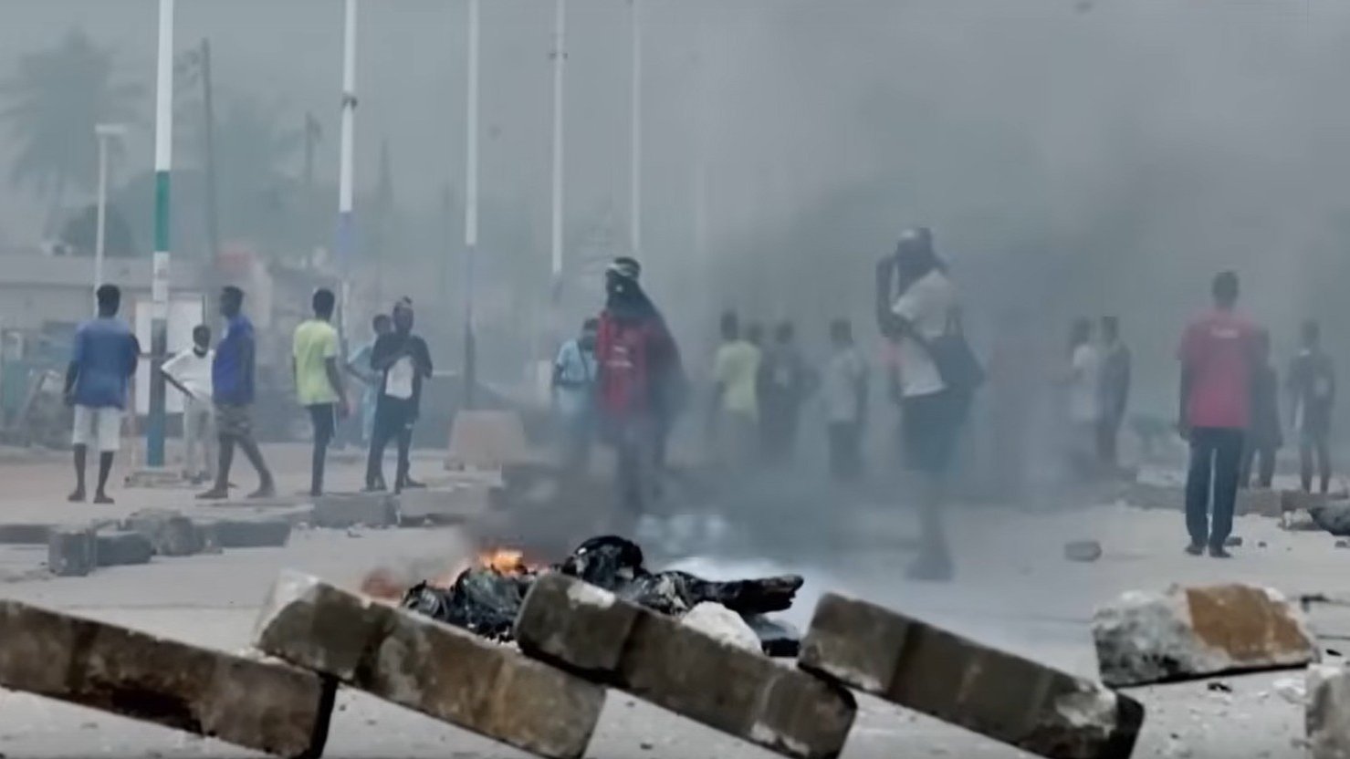 L'image montre une scène urbaine troublée. Au premier plan, plusieurs personnes courent rapidement, manifestant une impression d'urgence et de panique. En arrière-plan, de grands nuages de fumée noire s'élèvent dans l'air, indiquant un incendie. Des flammes orange et rouges jaillissent de barricades, suggérant une situation de conflit ou de désordre. Le décor urbain autour d'eux est flou, mais on peut discerner des bâtiments et des routes. L'atmosphère est tendue, évoquant un sentiment de danger et d'agitation.