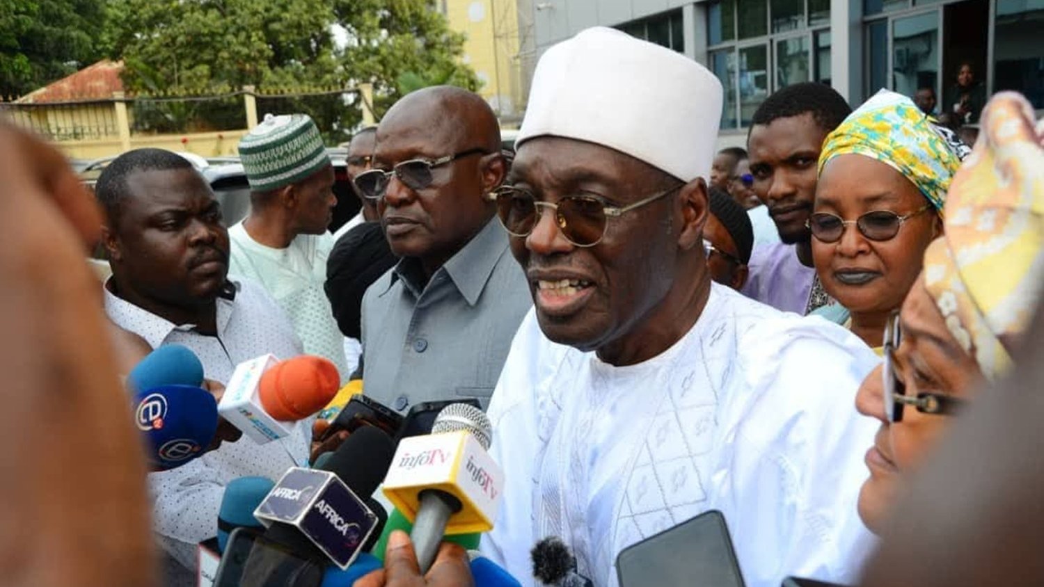Dans cette image, un groupe de personnes se trouve en train de discuter avec des journalistes. Au centre, un homme porte un costume traditionnel blanc, accompagné d'un chapeau traditionnel et d'une écharpe. Il parle aux médias, entouré de plusieurs personnes, dont certaines semblent prendre des notes ou poser des questions avec des micros devant eux. Les visages des personnes montrent un mélange d'intérêt et de concentration. En arrière-plan, on peut apercevoir un bâtiment moderne et des arbres, ce qui donne une ambiance urbaine à la scène.