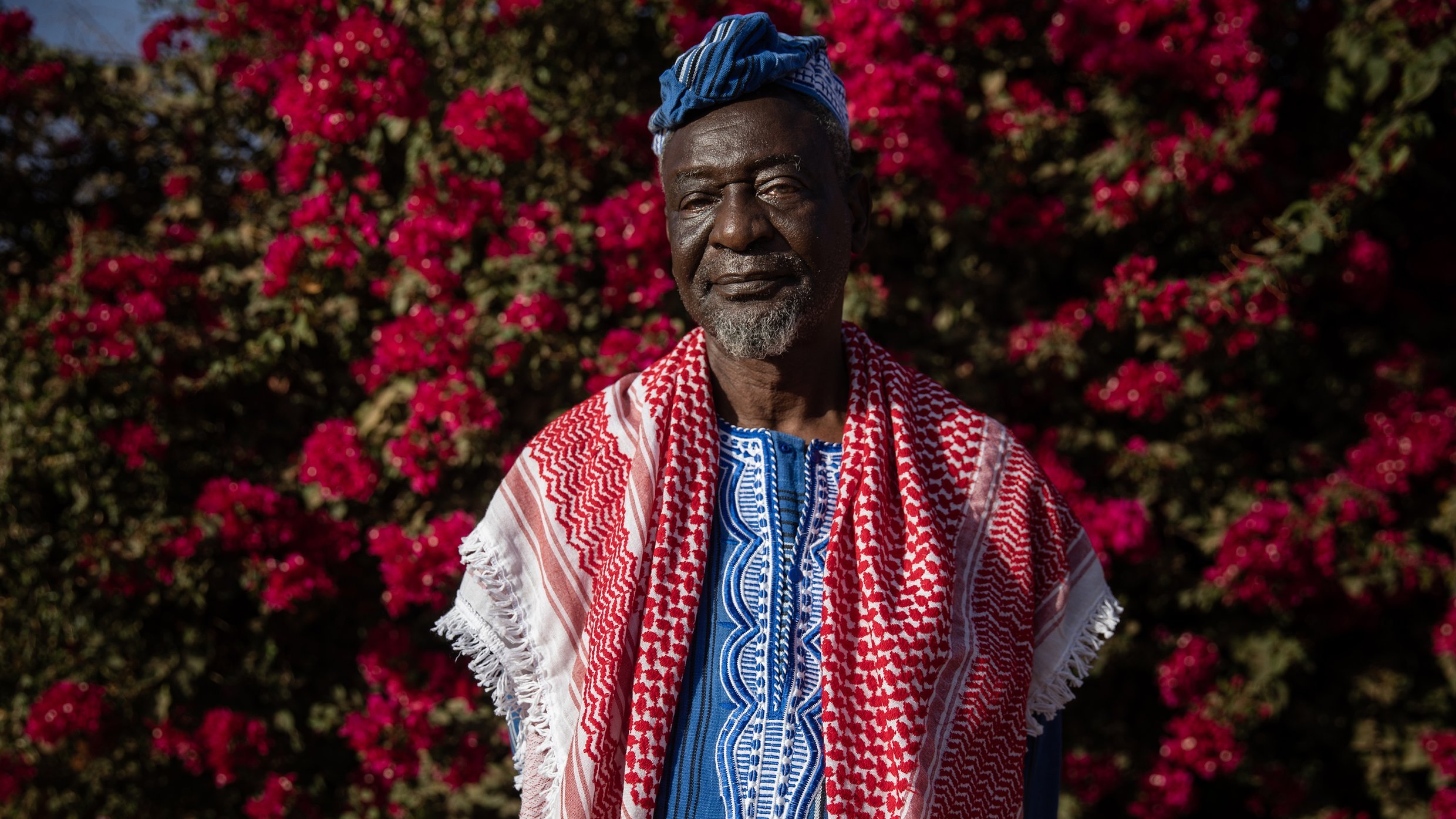 L'image montre un homme debout devant un fond vibrant de fleurs roses, probablement des bougainvilliers. Il porte une tenue traditionnelle riche en couleurs, avec un tissu bleu et des motifs complexes. Sur ses épaules, il a une écharpe ou un châle rouge et blanc, typique de certaines cultures. Son visage exprime sagesse et dignité, avec des traits marqués par le temps. Le contraste entre ses vêtements et les fleurs éclatantes crée une atmosphère chaleureuse et accueillante.