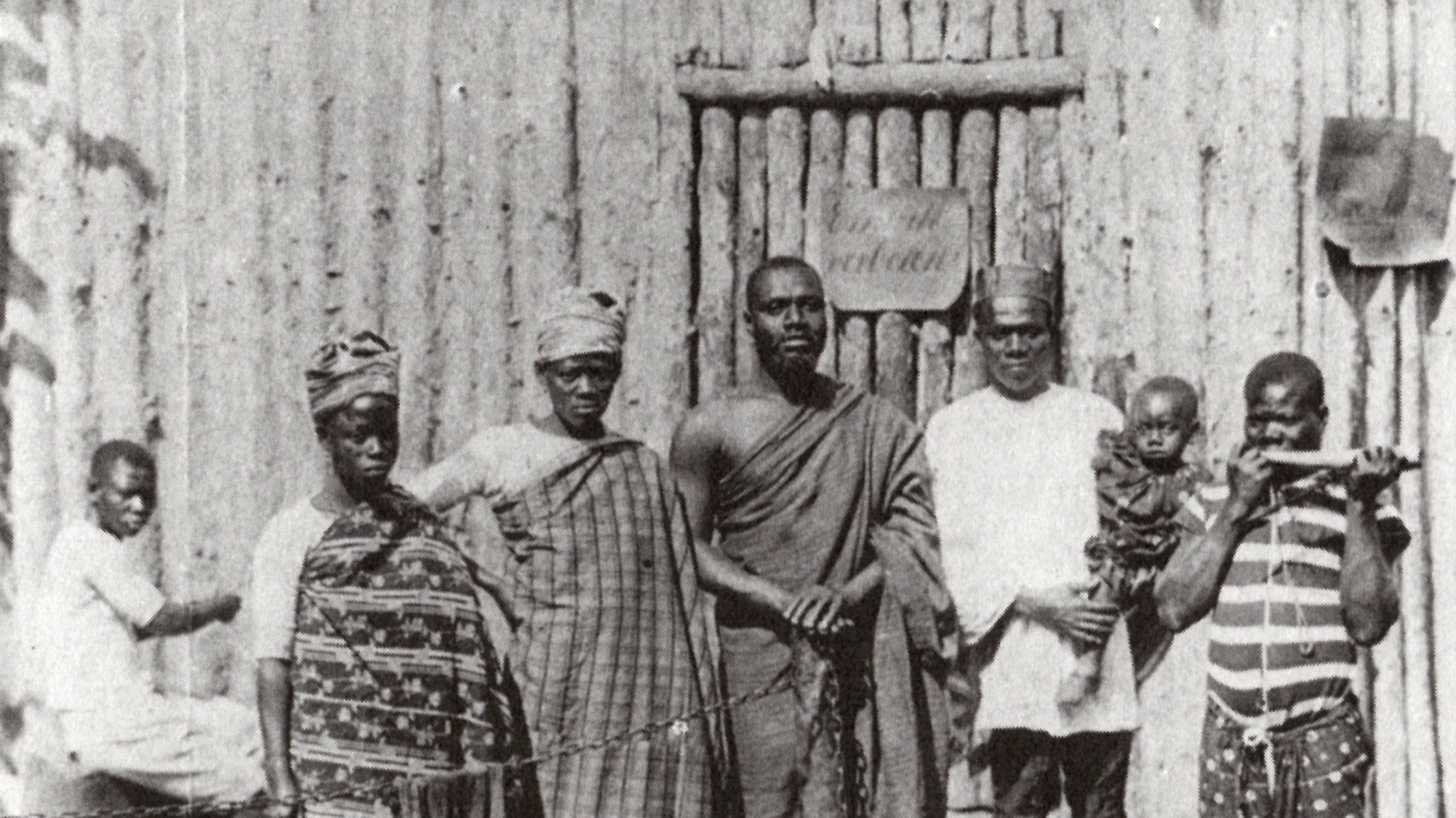 L'image montre un groupe de six personnes se tenant debout devant une structure en bois, qui semble être une maison ou un abri, avec un décor rustique. Au fond, on distingue un homme assis qui fait face à la scène principale. Les membres du groupe portent des vêtements traditionnels typiques, avec des motifs variés, affichant une richesse culturelle. L'un d'eux tient un enfant, tandis qu'un autre est équipé d'un bâton, suggérant une certaine autorité ou dignité. L'atmosphère de l'image dégage un sentiment de communauté et de convivialité.