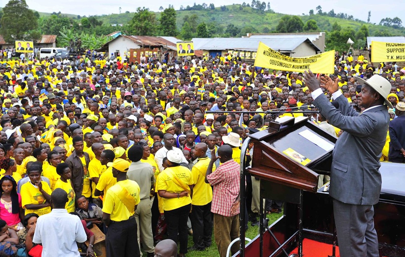 L'image montre une grande foule rassemblée en plein air. Les personnes dans la foule portent majoritairement des vêtements jaunes, ce qui pourrait indiquer qu'elles soutiennent un mouvement ou un parti spécifique. Au premier plan, un homme, vraisemblablement un orateur ou un dirigeant, se tient derrière un podium. Il lève les bras, semblant interagir avec la foule et exprimer un message puissant. En arrière-plan, on aperçoit des bâtiments simples et des collines verdoyantes, donnant un cadre naturel à l'événement. L'atmosphère semble énergique et mobilisée.