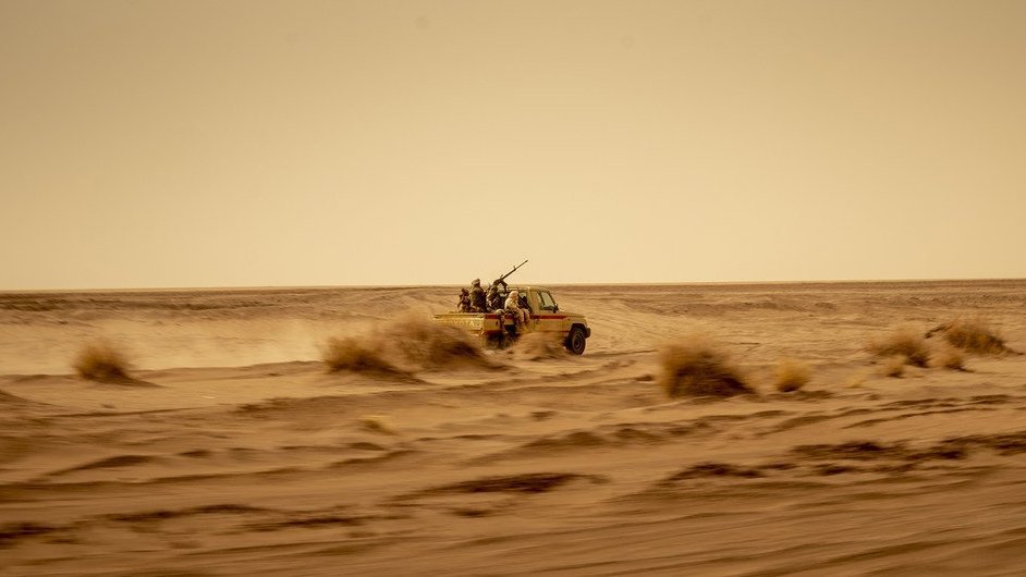 L'image montre un véhicule tout-terrain, probablement un pick-up, qui avance rapidement à travers un désert. La poussière s'élève autour de lui, créant une ambiance de mouvement et de vitesse. À l'arrière du véhicule, on aperçoit des silhouettes de personnes, probablement des passagers, qui semblent être équipés pour un environnement hostile. Le paysage est vaste et aride, avec des dunes de sable et des buissons résistant aux conditions sèches. Le ciel est clair mais teinté d'une couleur jaunâtre, suggérant une chaleur intense. L'ensemble donne une impression de solitude et d'aventure dans un milieu difficile.