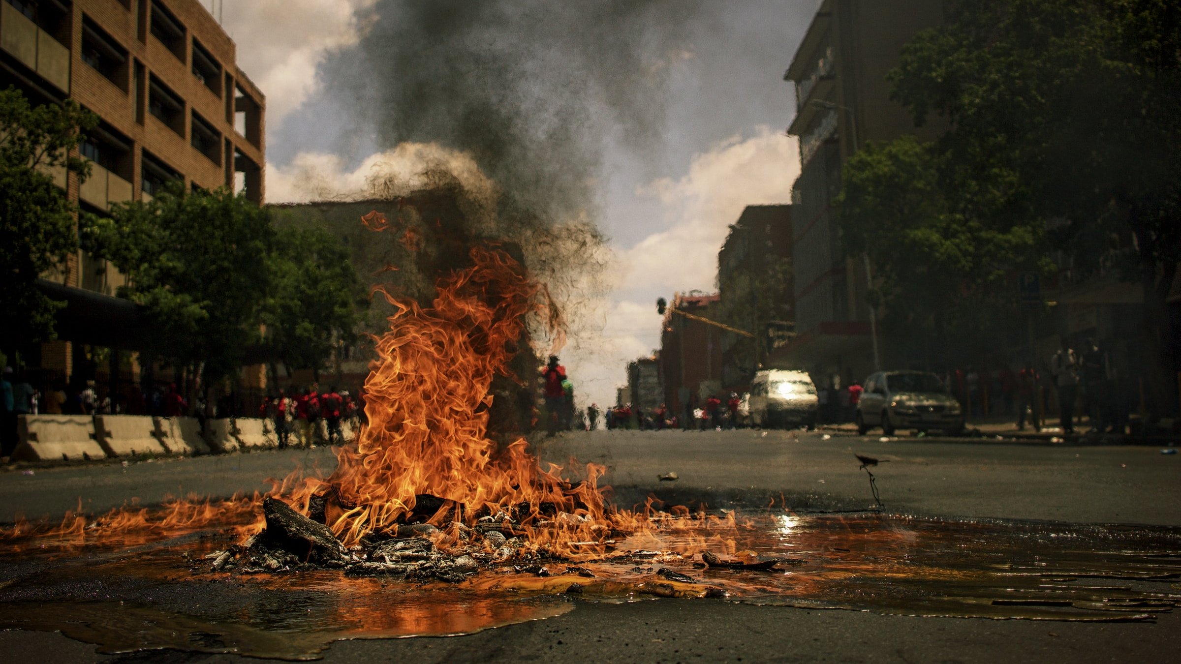 L'image montre une rue animée où un feu brûle au centre. Des flammes vives et tourbillonnantes s'élèvent de ce qui semble être des débris sur le sol. Une épaisse fumée noire s'échappe vers le ciel, obscurcissant partiellement la lumière du jour. On peut apercevoir des bâtiments sur les côtés de la rue, ainsi qu'un groupe de personnes au fond, créant une atmosphère tendue et dynamique. L'air est probablement chargé d'odeurs de brûlé, et le son du crépitement du feu se mêle à une ambiance vibrante de manifestation ou d'agitation.