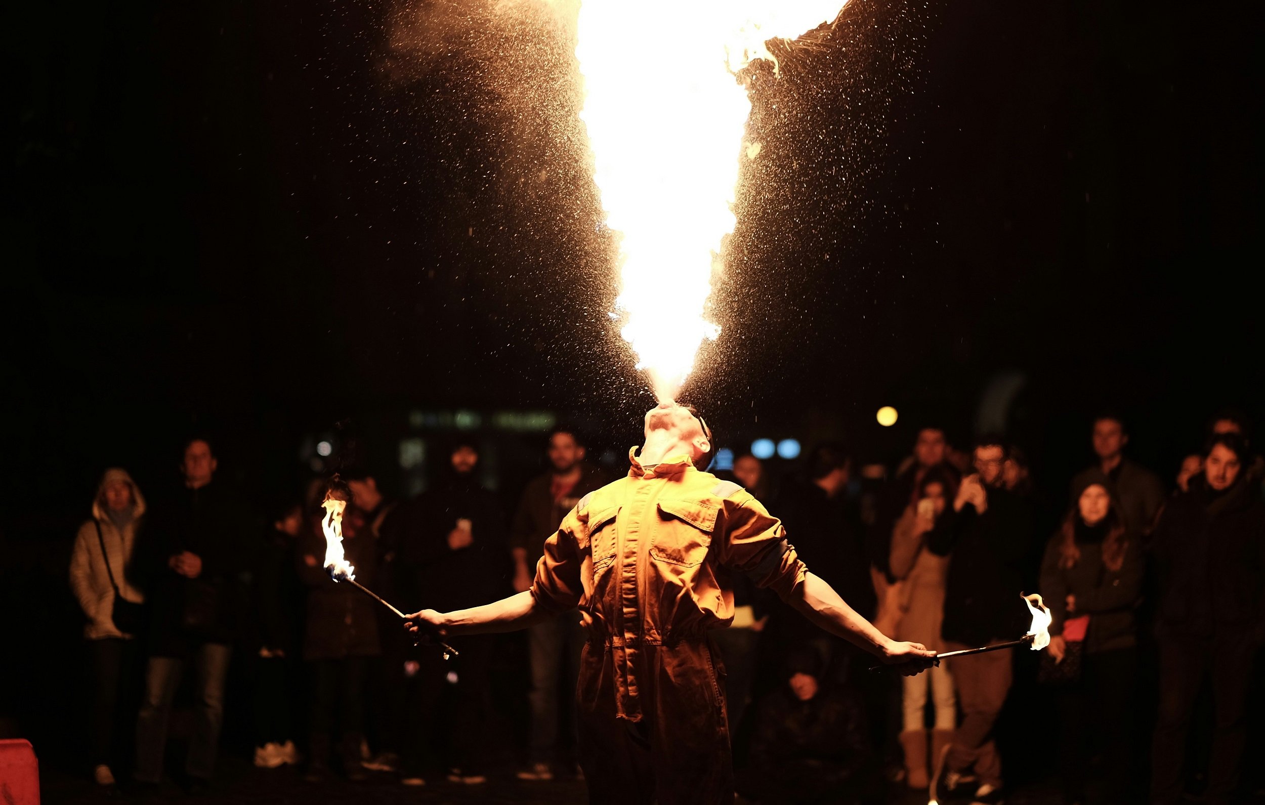 L'image présente un artiste de rue en action, exécutant un numéro de jonglage avec du feu. Il se tient au centre de la scène, le dos arqué et la tête inclinée en arrière, tandis qu'il crache une immense flamme qui s'élève vers le ciel. L'artiste porte un costume de couleur sombre, et ses bras sont tendus, chacun tenant un bâton de feu. En arrière-plan, une foule captivée et attentive observe la performance, créée par la magie du feu illuminant la nuit. L'atmosphère est électrique, mêlant curiosité et émerveillement face à ce spectacle audacieux.