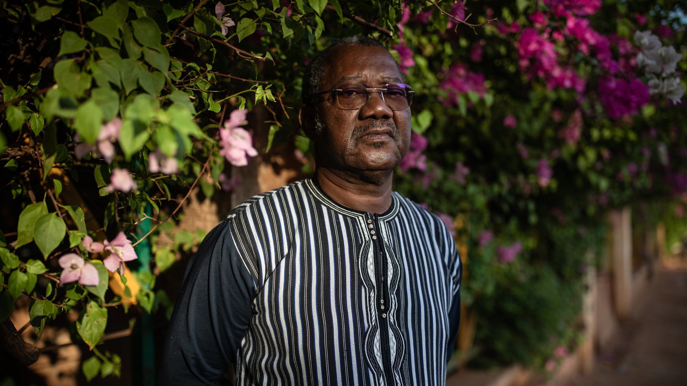 L'image montre un homme debout devant un mur recouvert de magnifiques bougainvilliers aux couleurs vives, principalement en rose et violet. Il porte une chemise à rayures noires et blanches, avec un col qui attire l'attention. Son expression est sérieuse et réfléchie, et il semble absorbé par ses pensées. En arrière-plan, on peut percevoir une ambiance douce et chaleureuse grâce à la lumière naturelle qui illumine la scène, accentuant la beauté des fleurs.