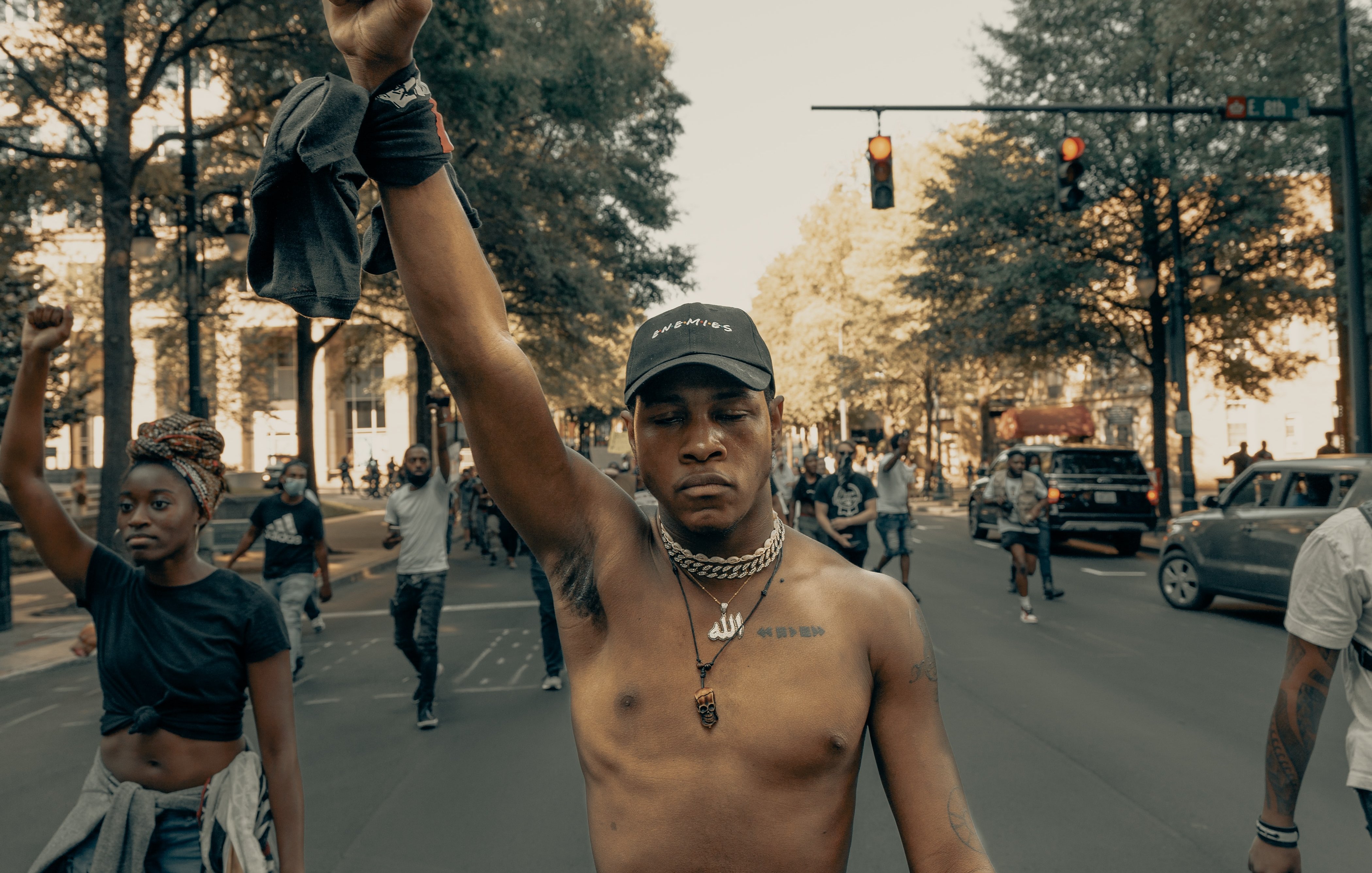 L'image montre une scène de manifestation dans une rue. Au premier plan, un jeune homme, torse nu, lève le poing en signe de protestation. Il porte une casquette et des chaînes autour du cou. À l'arrière-plan, d'autres personnes marchent, certaines levant également le poing, témoignant d'un esprit collectif. La rue est bordée d'arbres, et on peut apercevoir des voitures et des feux de signalisation. L'ambiance semble énergique et engagée, reflétant une lutte pour la justice ou l'égalité.