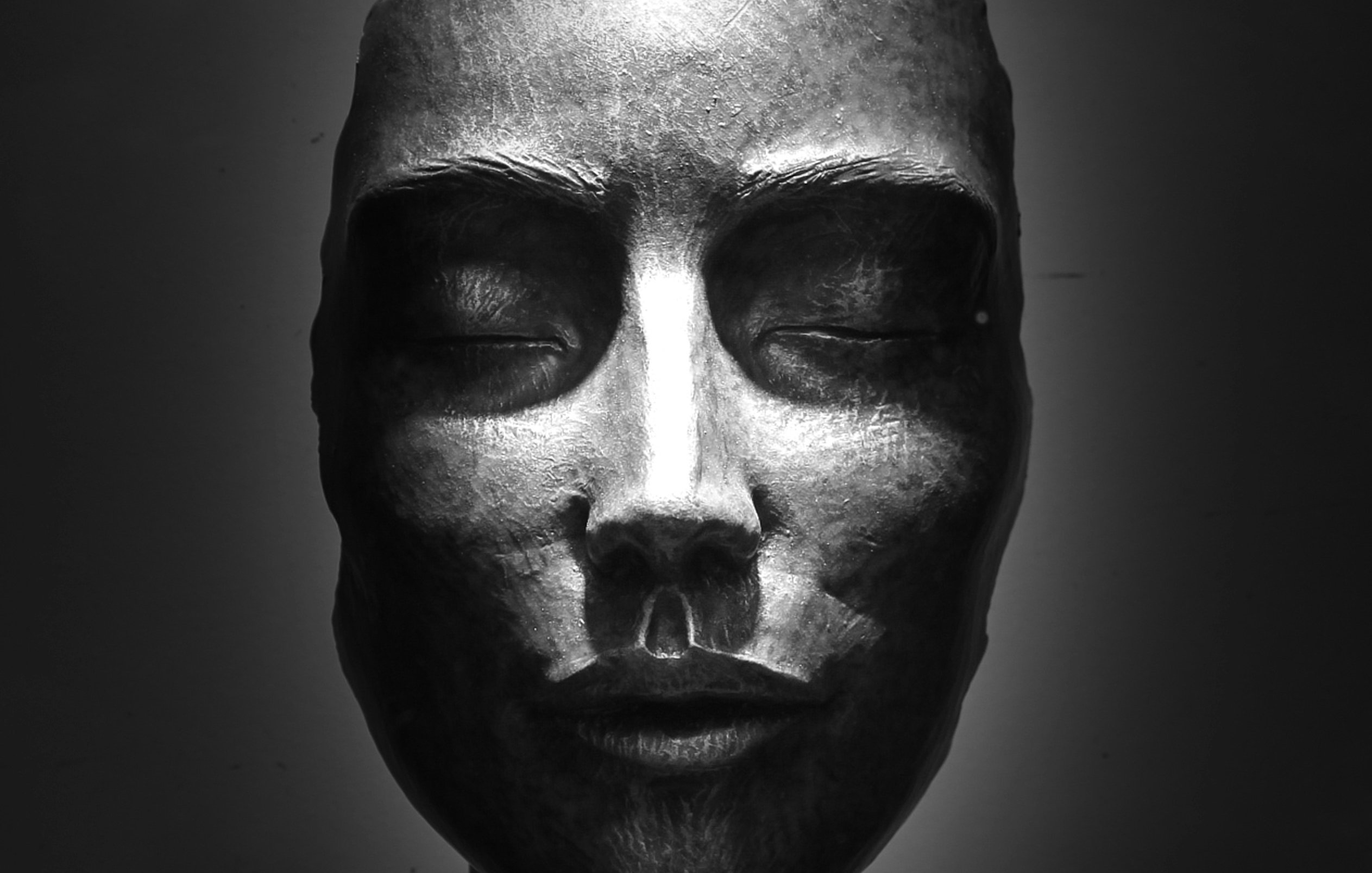 L'image représente une sculpture en bronze d'un visage humain. Celui-ci est stylisé et dégage une expression de sérénité. Les yeux sont fermés, et les traits sont à la fois doux et anguleux. La lumière tombe subtilement sur le visage, créant des ombres qui accentuent les contours de la structure. La sculpture est posée sur une base solide, et la posture semble évoquer une sensation de calme et de réflexion. Des détails comme le nez et les lèvres sont finement sculptés, ajoutant une profondeur à l'œuvre. L'ensemble évoque un sentiment de paix intérieure.