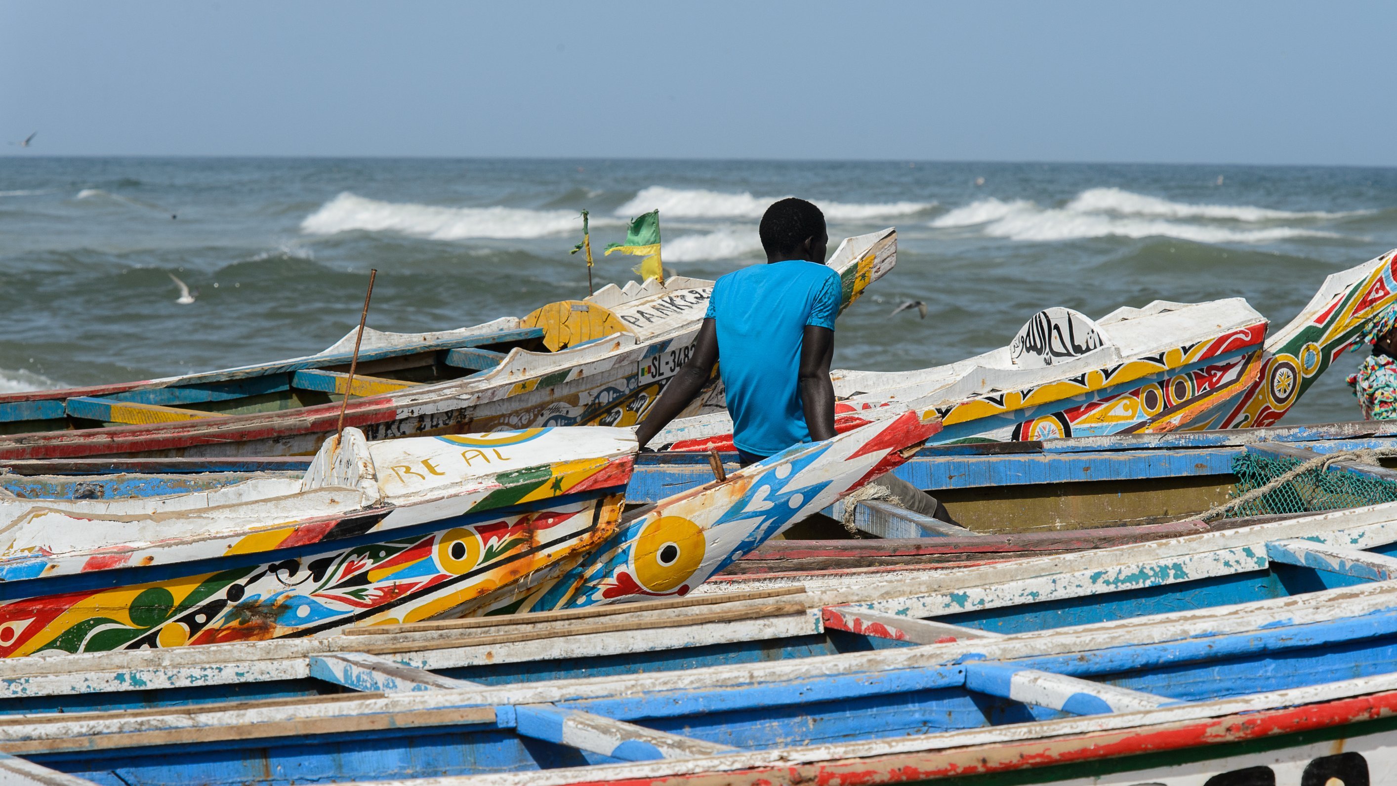 L'image montre une plage animée au bord de la mer. Des bateaux colorés se chevauchent sur le sable, avec des motifs vifs et des teintes variées, tels que le bleu, le rouge et le jaune. Un homme, vêtu d'un t-shirt bleu, est assis au bord des bateaux, tourné vers l'océan. Les vagues s'écrasent doucement sur le rivage, créant un bruit apaisant. Dans l'air, on peut sentir une brise marine fraîche et éventuellement entendre le cri des oiseaux qui volent autour. L'ambiance dégage une sensation de tranquillité tout en étant dynamique grâce à la présence des bateaux et de l'homme observant l'horizon.