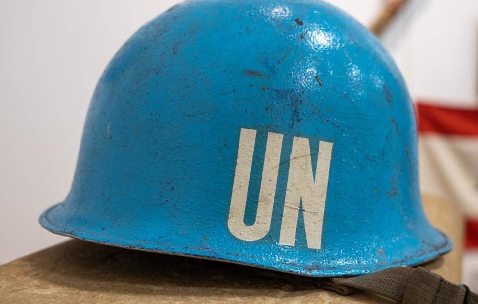L'image montre un casque de couleur bleu vif, typique des casques de l'ONU. La surface du casque est légèrement abîmée, indiquant qu'il a probablement été utilisé sur le terrain. Sur le devant, des lettres blanches "UN" sont bien visibles, représentant l'Organisation des Nations Unies. Le casque est posé sur une surface plane, avec un fond flou qui évoque un environnement de musée ou d'exposition, où des objets historiques peuvent être présentés. Ce casque symbolise la paix et les missions de maintien de la paix menées par l'ONU dans diverses régions du monde.