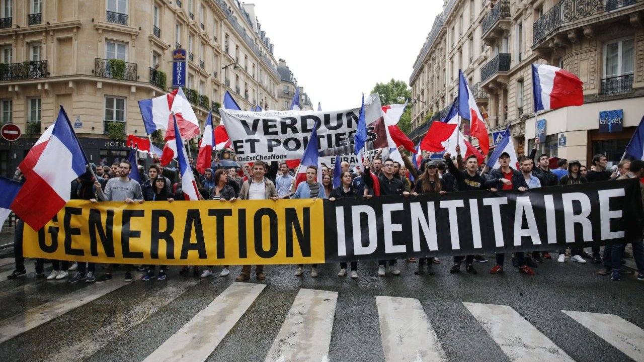 L'image montre une manifestation dans une rue urbaine. Un groupe de personnes tient une grande banderole noire avec le texte "GENERATION IDENTITAIRE" en lettres jaunes bien visibles. Elles brandissent également de nombreux drapeaux tricolores français, qui flottent au-dessus d'elles. En arrière-plan, on aperçoit une autre bannière avec les mots "VERDUN ET POITERS", indiquant probablement des messages ou des revendications relatives à ces lieux. Les manifestants semblent unis et déterminés, marchant ensemble sur le passage piéton, tandis que des bâtiments typiques de la ville se dressent à leurs côtés. L'ambiance générale est celle d'une démonstration publique, avec une forte présence de symboles nationaux.
