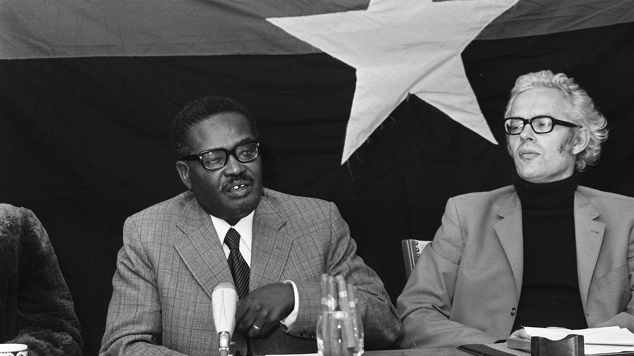 L'image montre une réunion où deux hommes sont assis à une table. À gauche, un homme d'âge moyen, portant des lunettes et un costume gris à carreaux, semble s'adresser aux autres participants, gesticulant légèrement avec une main. Il a une expression attentive et engagée. À sa droite, un homme plus âgé avec des cheveux gris et des lunettes, habillé d'un pull noir et d'une veste beige, l'écoute avec intérêt. En arrière-plan, on devine un grand drapeau noir avec une étoile blanche, ajoutant un contexte politique à la scène. Sur la table, il y a quelques verres, un microphone et un bloc-notes. L'atmosphère semble sérieuse et concentrée.