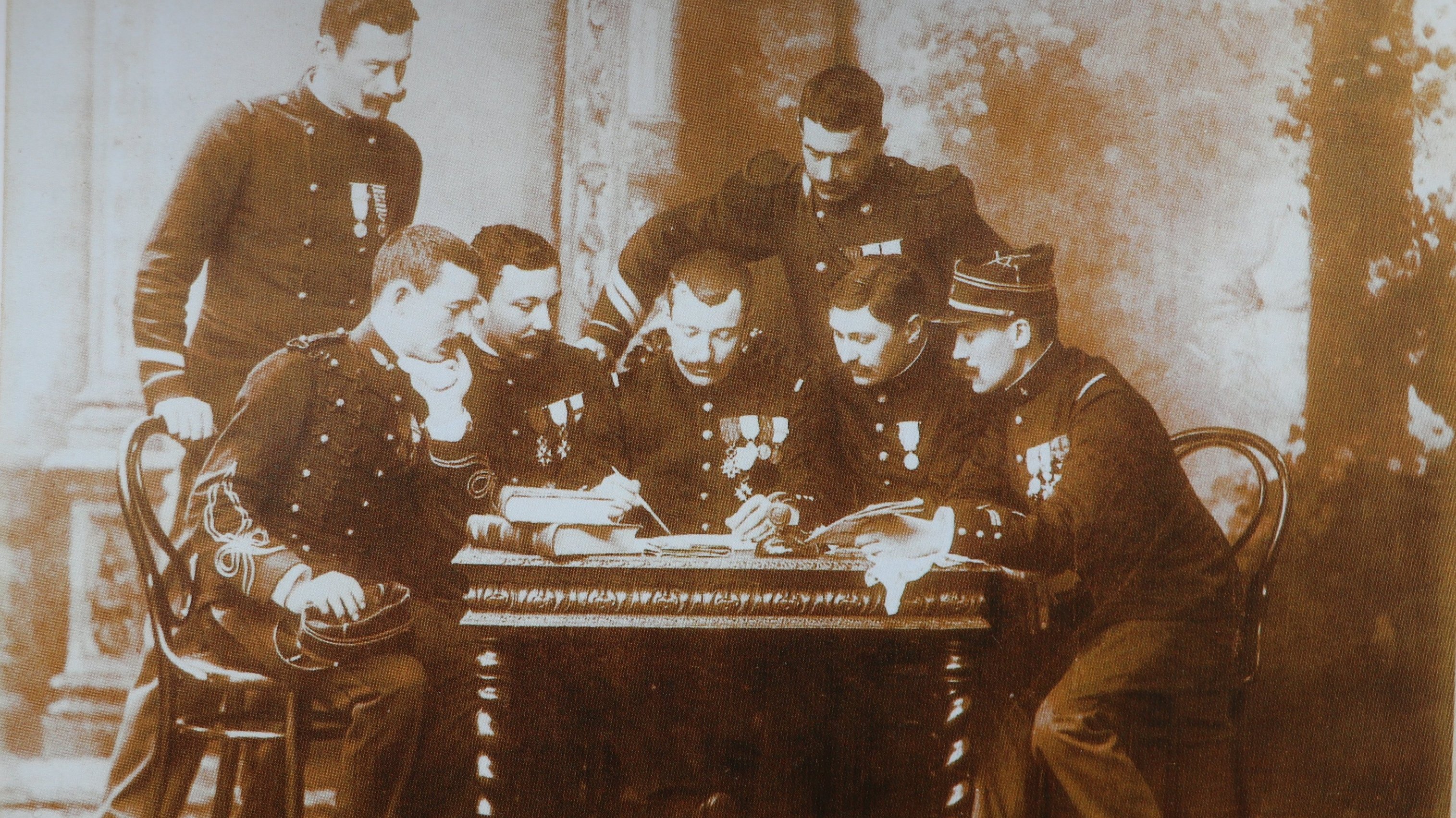 Cette image en noir et blanc représente un groupe de six hommes en uniforme, assis autour d'une table en bois. Ils semblent être des membres d'un corps militaire, chacun portant des médailles décoratives sur leur uniforme. Les hommes sont concentrés sur un document ou un plan posé sur la table, et leur posture indique un échange d'idées ou de discussions sérieuses. En arrière-plan, on peut distinguer quelques éléments architecturaux flous, suggérant un intérieur de bâtiment classique. L'atmosphère se dégage une certaine solennité et camaraderie.