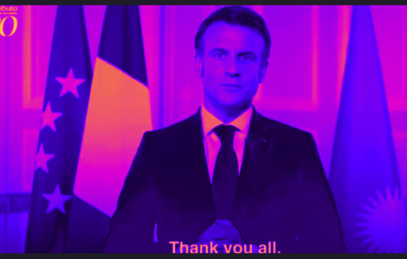 L'image montre un homme en costume sombre, debout devant un arrière-plan aux tons violets et roses. Il a une expression sérieuse et est placé entre deux drapeaux : un drapeau tricolore français à gauche et un autre qui pourrait représenter l'Argentine à droite. Ses mains sont jointes devant lui, comme s'il allait s'exprimer. En bas de l'image, on peut lire un texte en anglais qui dit "Thank you all", indiquant qu'il s'adresse à un auditoire. L'ensemble de la scène dégage une atmosphère formelle et solennelle.