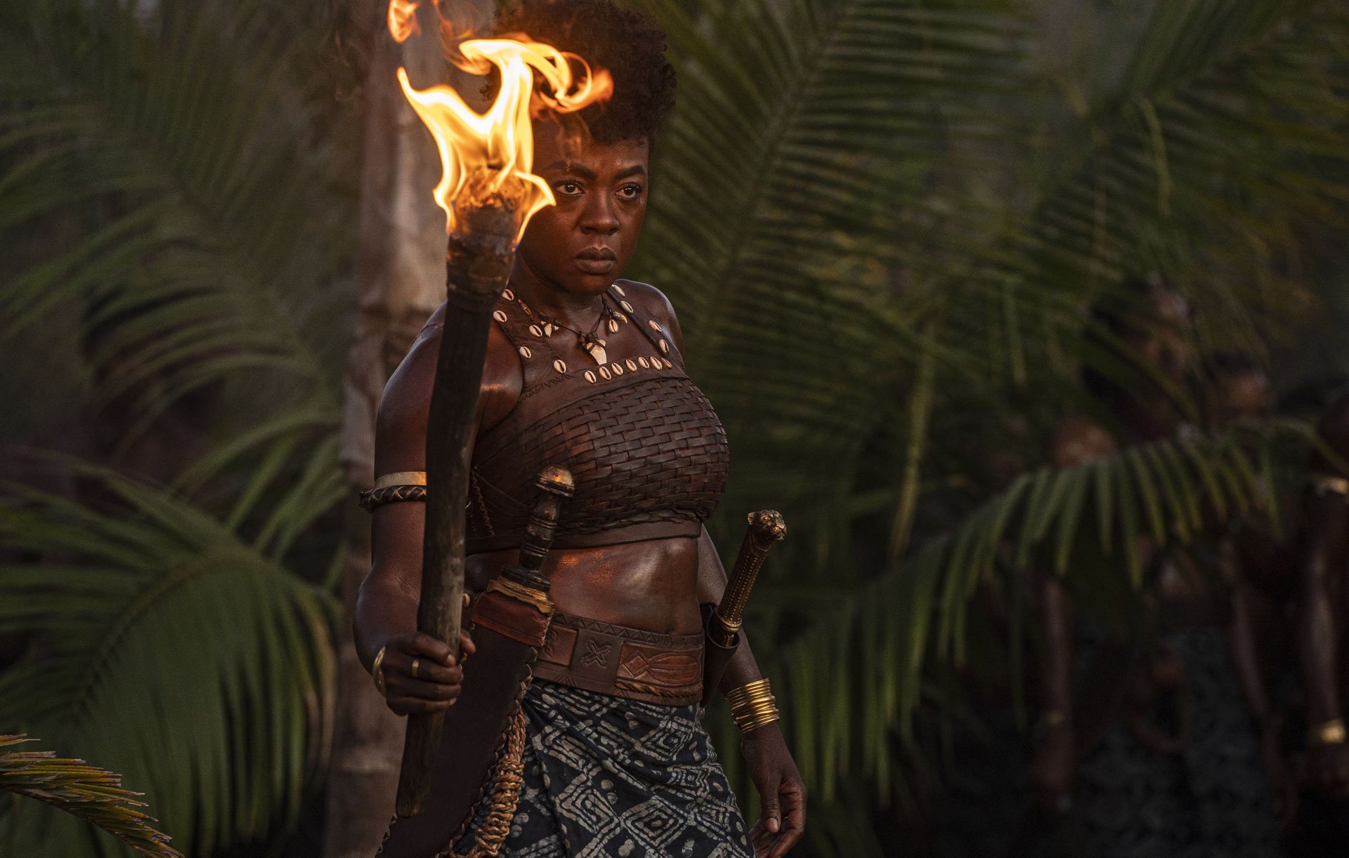 L'image montre une femme debout au milieu d'une luxuriante végétation tropicale, avec des palmiers en arrière-plan. Elle tient une torche enflammée dans sa main droite, créant une lumière chaleureuse qui éclaire son visage déterminé. Elle porte un haut traditionnel en cuir et une jupe décorée de motifs. Des bracelets dorés ornent ses bras, ajoutant une touche d'élégance à son apparence. Son expression est forte et intense, reflétant à la fois courage et détermination. L'atmosphère de l'image évoque une ambiance de puissance et de leadership.