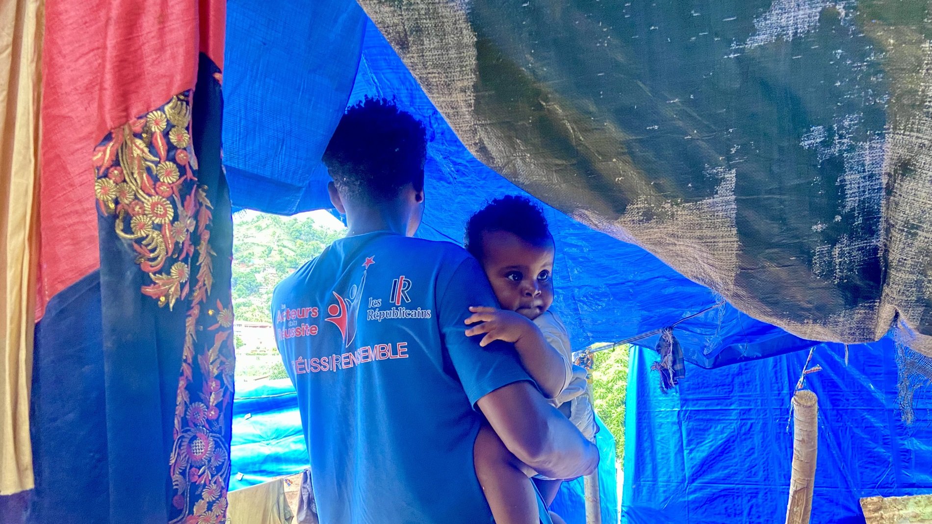 L'image montre un homme portant un enfant dans ses bras. Ils se trouvent sous une tente improvisée faite de tissus aux couleurs vives comme le bleu et le rouge. Le décor semble rural, avec des éléments naturels en arrière-plan. L'homme a un t-shirt bleu avec des inscriptions, et l'enfant, qui regarde légèrement en arrière, semble être de très jeune âge. La scène évoque un moment de tendresse et de protection dans un environnement simple.