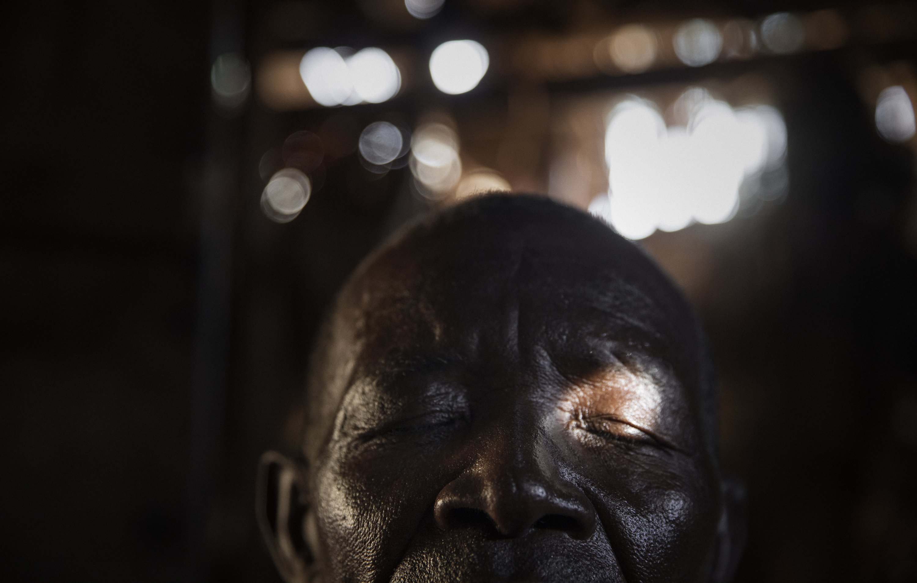 L'image montre un homme avec les yeux fermés, laissant transparaître une profonde sérénité. Son visage, marqué par le temps, présente des rides et une peau sombre. Une lumière douce filtre à travers un espace, touchant délicatement son visage et créant des reflets subtilement brillants en arrière-plan. L'ambiance de l'image est calme, évoquant un moment de méditation ou de paix intérieure. Les contours flous de l'environnement suggèrent un espace simple, sans détails distracteurs.